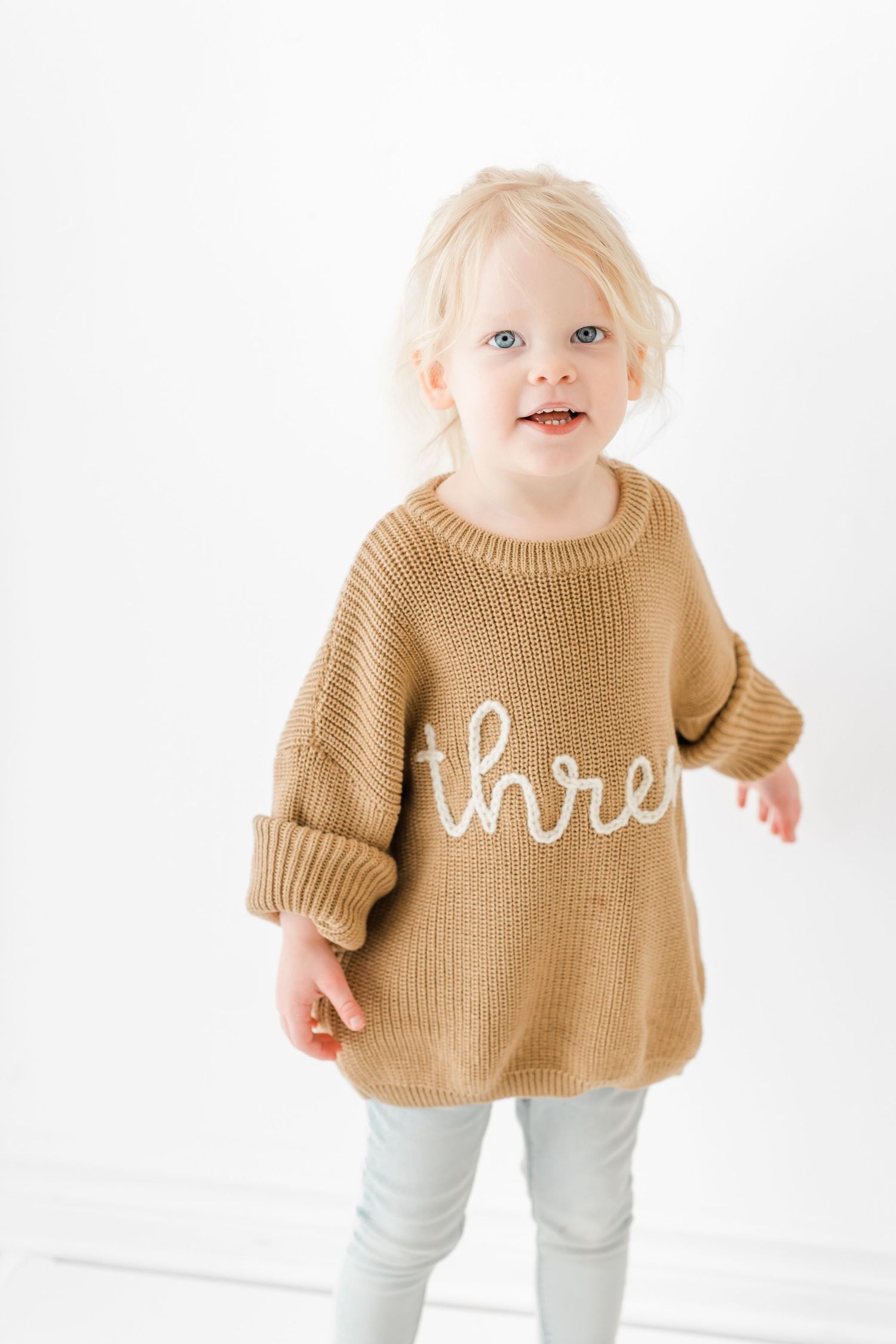 Small child wearing a cozy tan knit sweater with white text and light colored pants against white background.