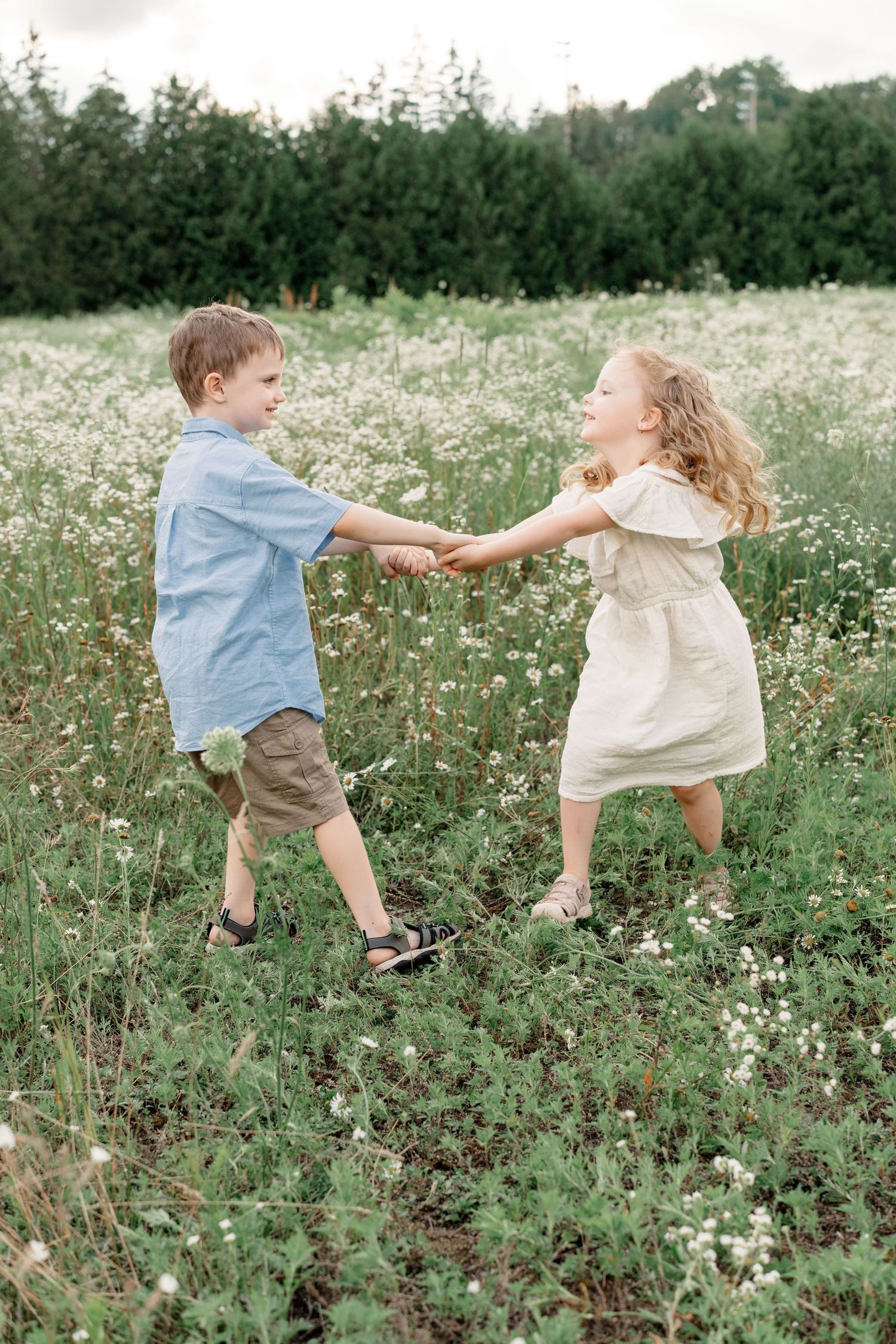 Two children dance and play together in a summer meadow filled with delicate white wildflowers and green grass.