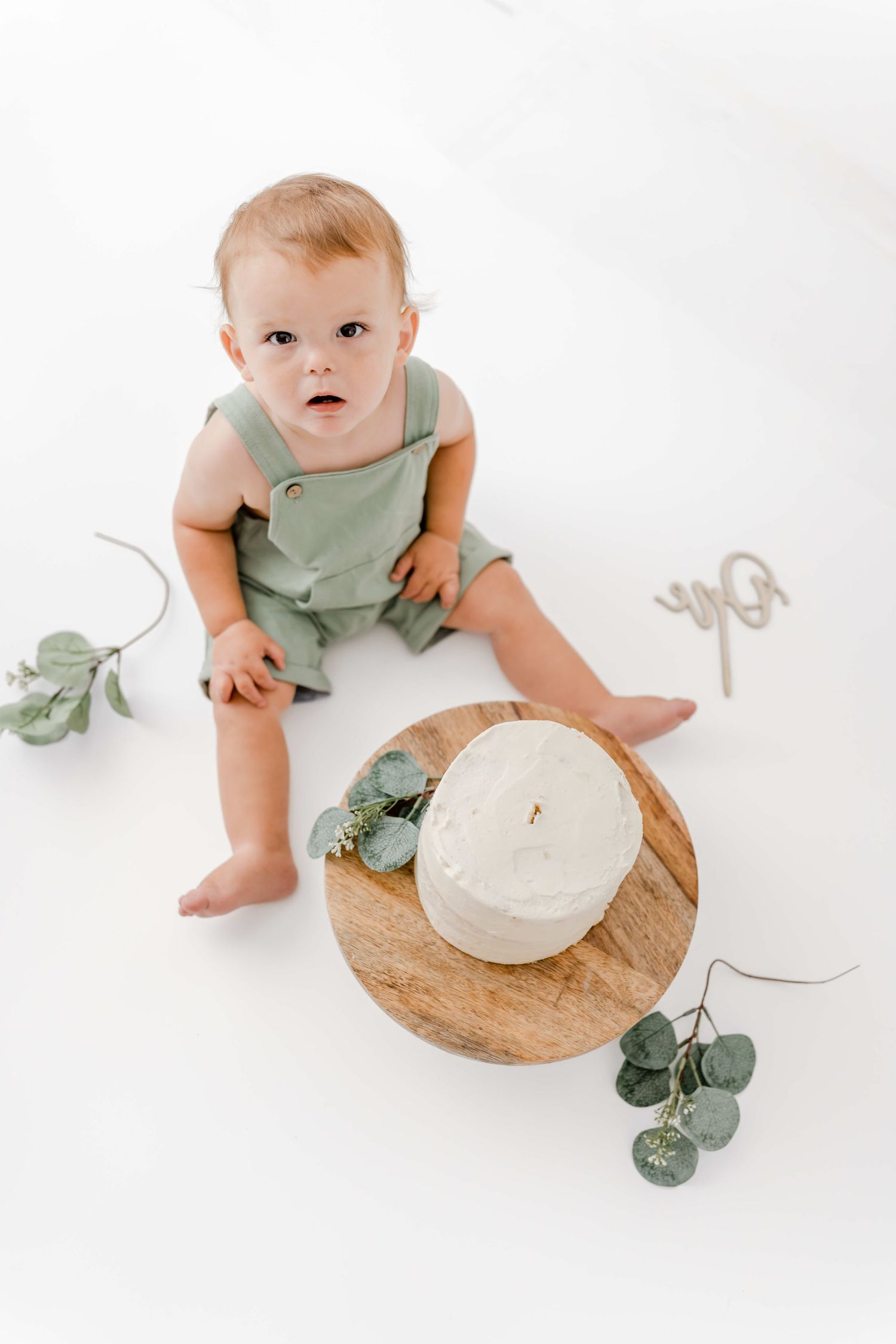 Baby in sage green romper sits near white pumpkin and eucalyptus on wooden board in minimalist photo series.