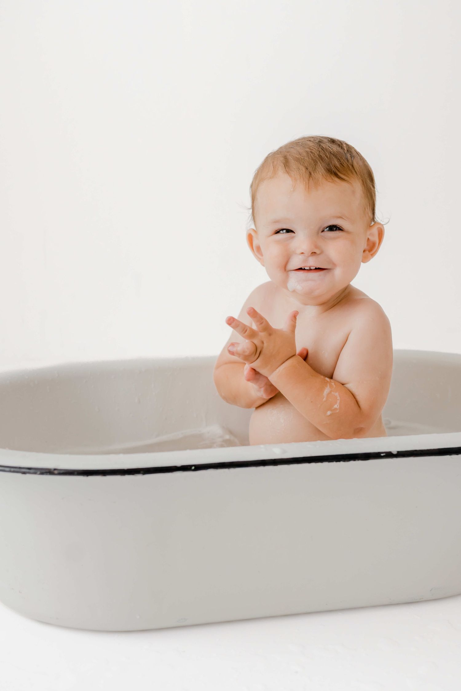 A happy baby sits in a minimalist white bathtub during bath time, smiling and playing.