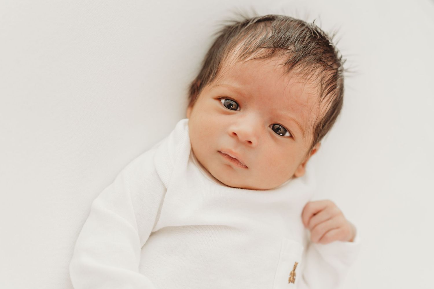 Newborn baby lying on white blanket with peaceful expression and dark hair.