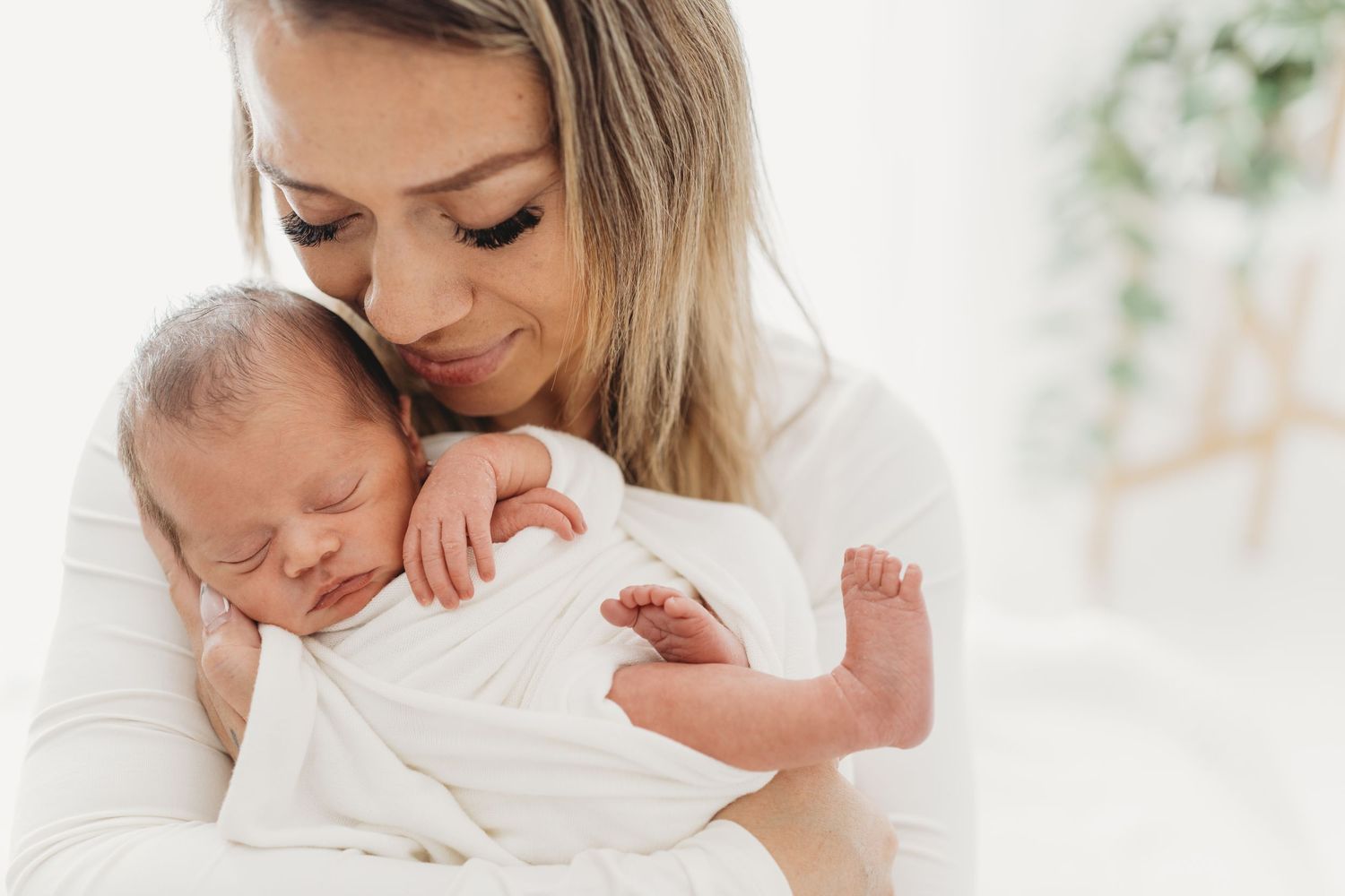 A tender moment between parent and newborn baby in soft white tones with greenery in the background.