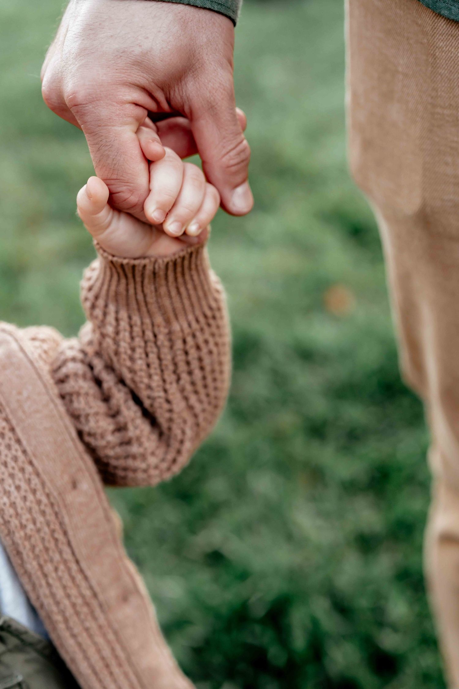 Close-up view of a small child's hand holding an adult's finger while wearing a knit sweater outdoors.