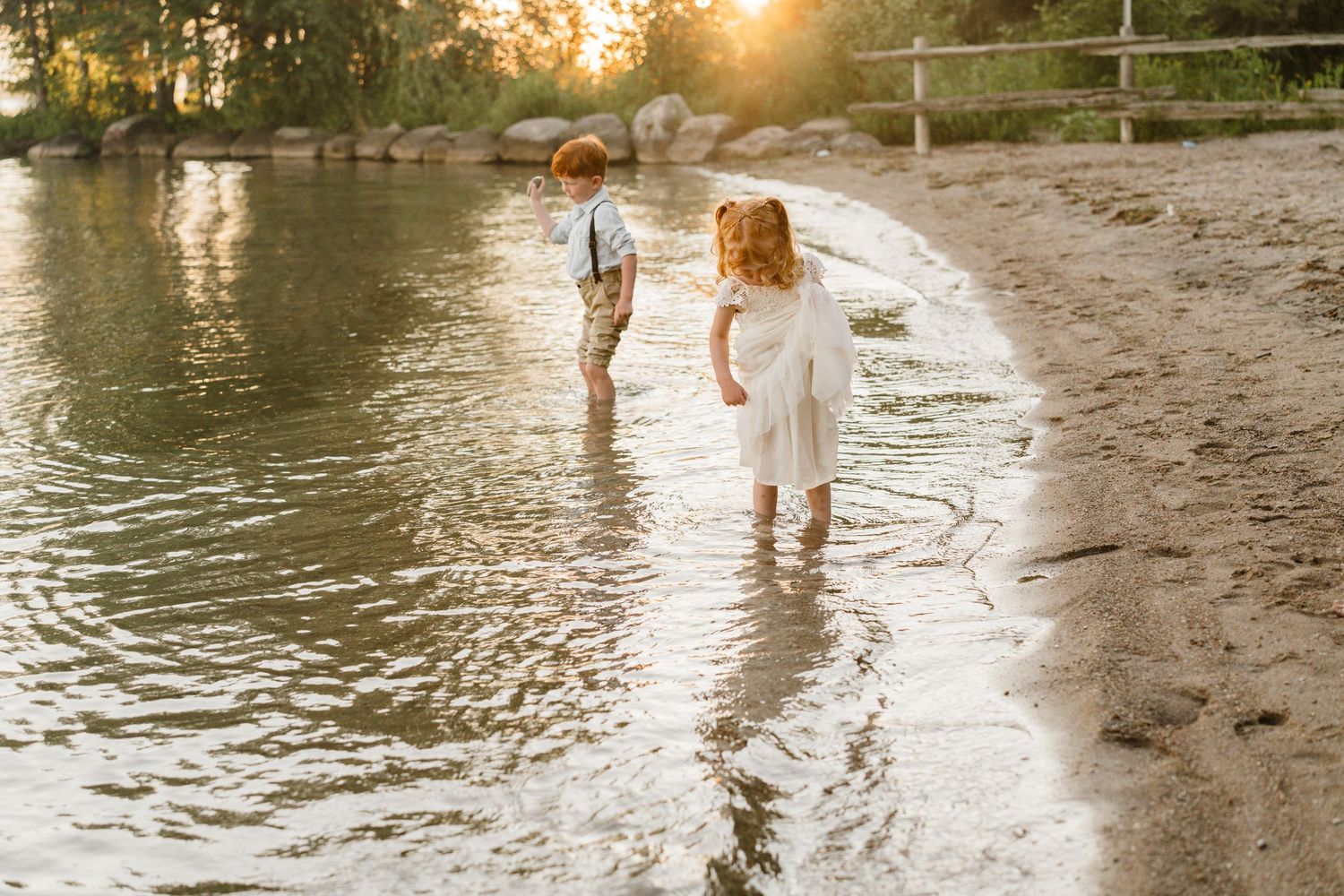Two young children wade in shallow water along a sandy shoreline during a golden sunset.
