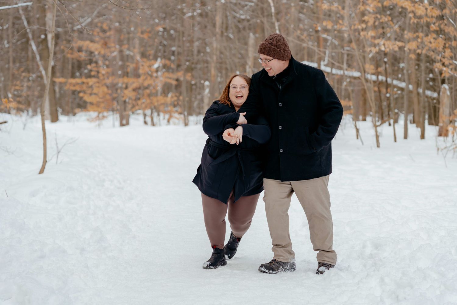 A couple walks together on a snowy path through a winter forest, both wearing warm black coats.