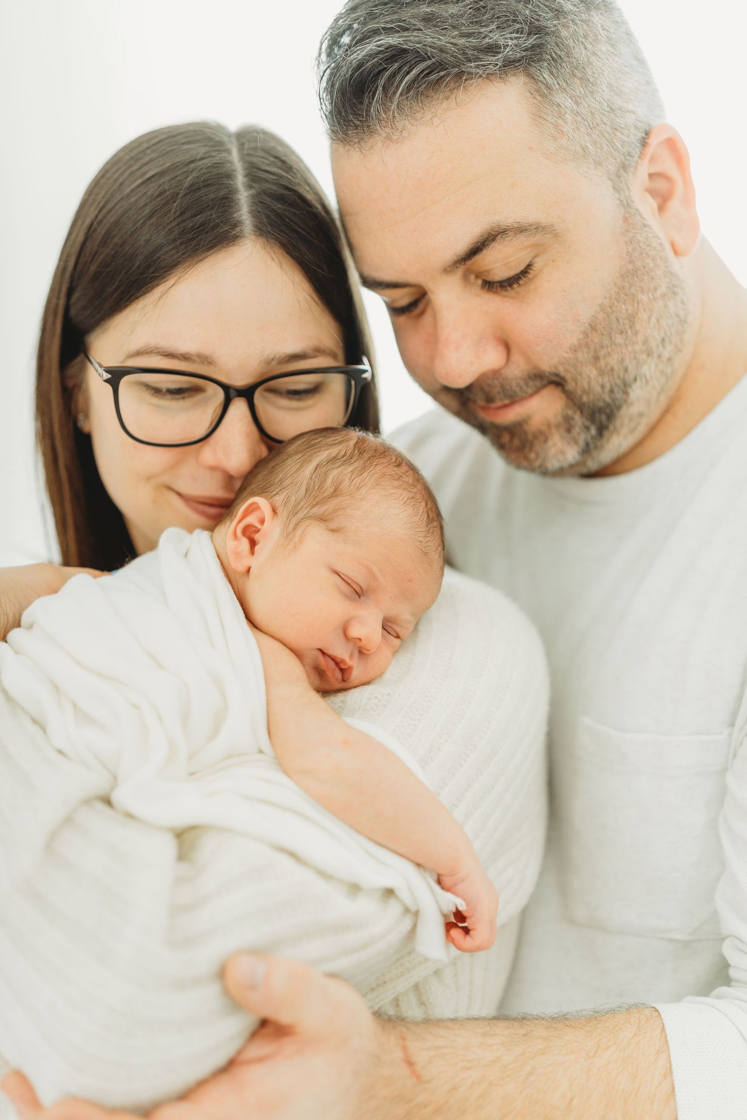 A couple in white clothing holds and gazes lovingly at their sleeping newborn baby in a bright, minimalist setting.