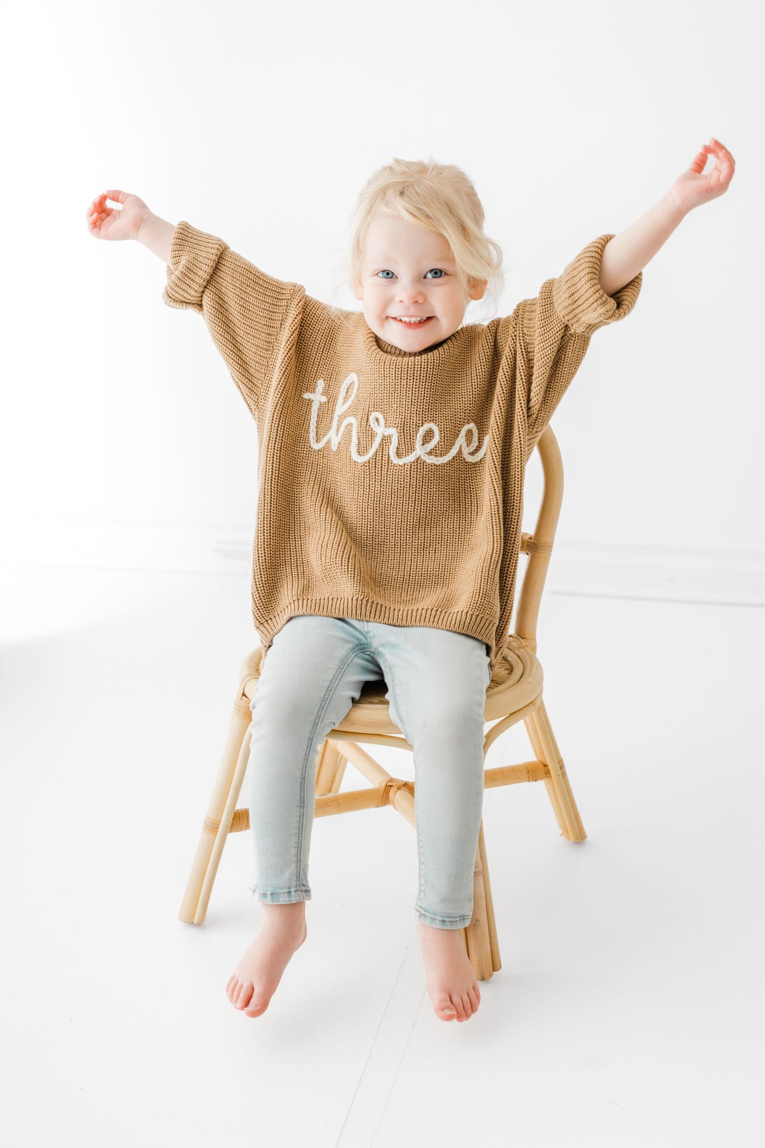 Child wearing tan sweater with 'three' text sits on wooden chair with arms raised in celebration against white background.