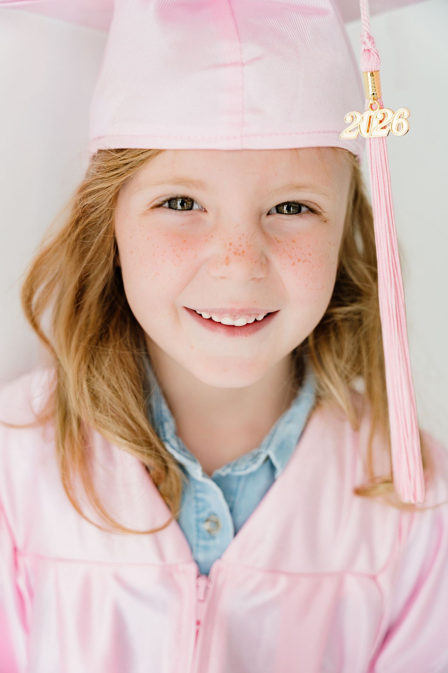 Young girl smiling in pink graduation cap and gown with 2026 tassel charm, wearing a denim shirt underneath.