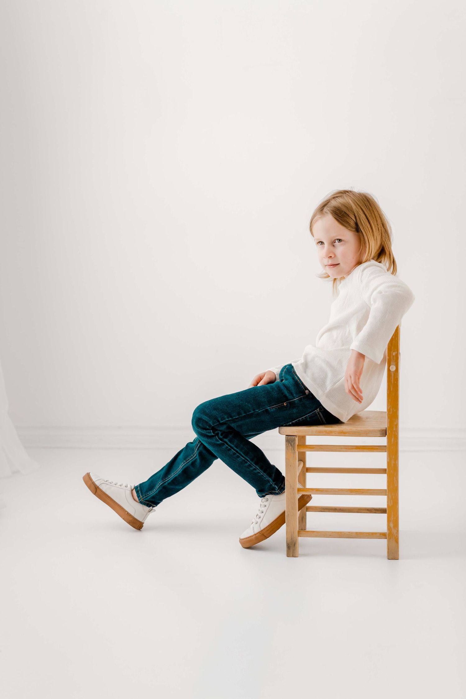 Child in white shirt and blue pants performs playful sequence of poses on wooden chair against white backdrop.