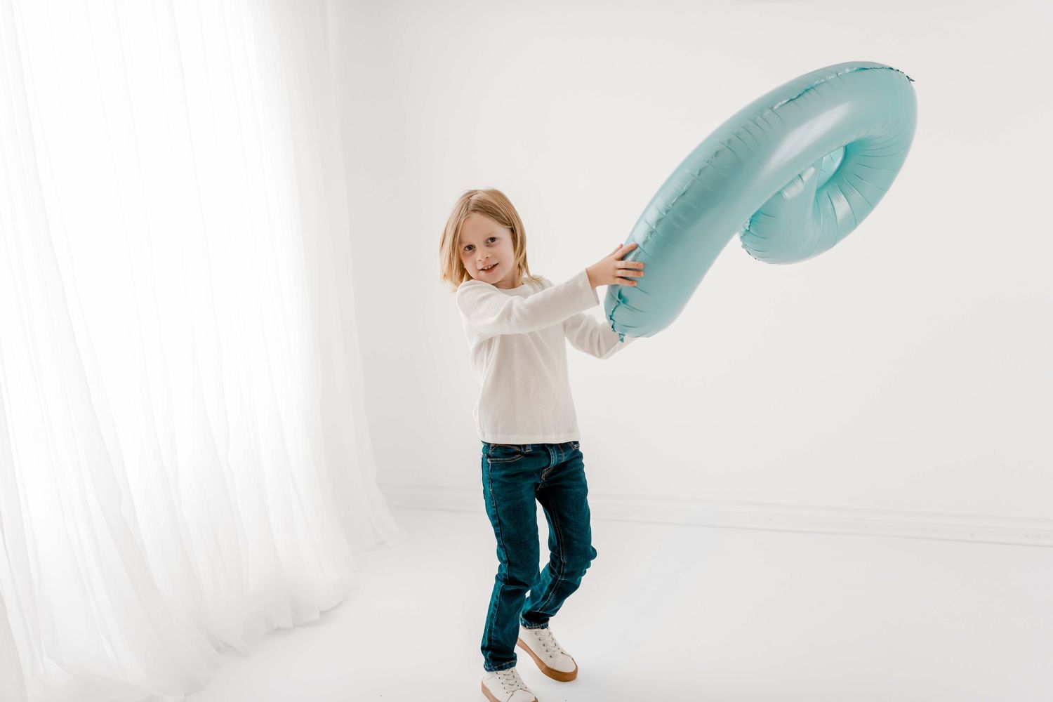 Child in white sweater and jeans playfully waves turquoise fabric in bright white studio setting.