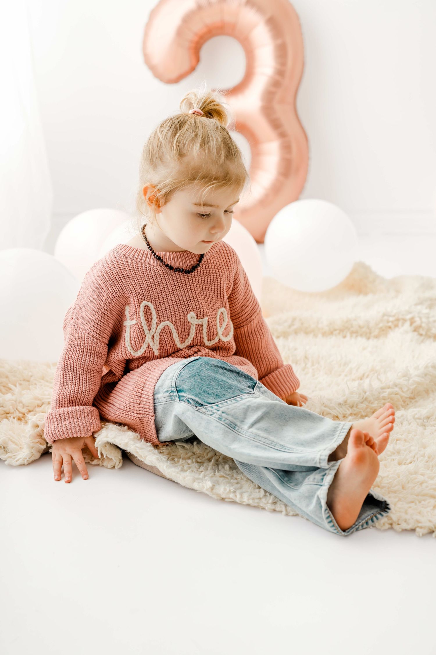 A child in a pink sweater with 'three' text and jeans sits on a white surface with rose gold number 3 balloon decor.