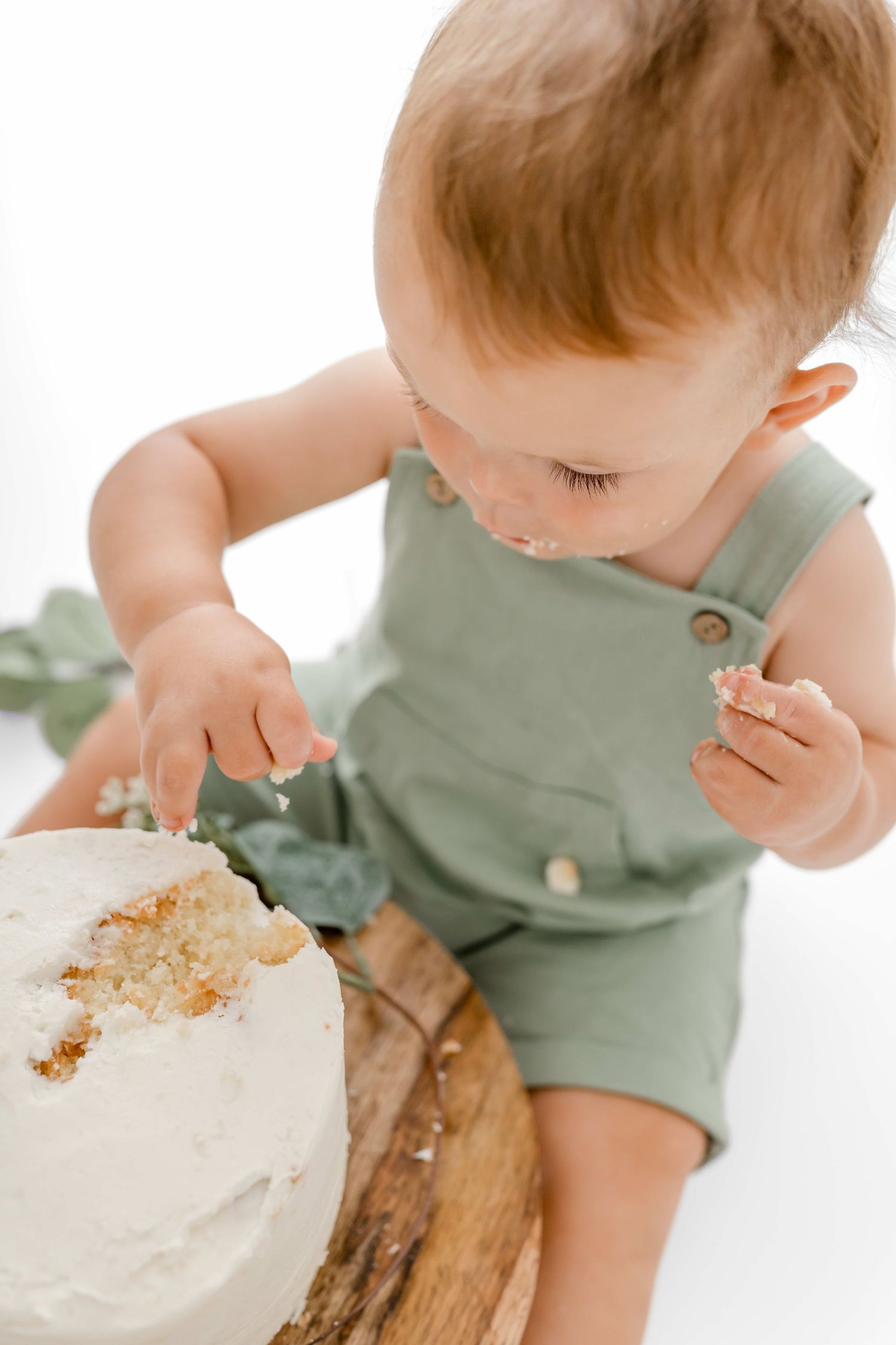 Baby in sage green romper explores and plays with white frosted cake during cake smash photo session.