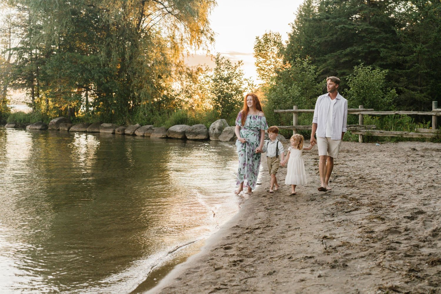 Family of four walking along a sandy riverbank at golden hour, surrounded by lush green trees.