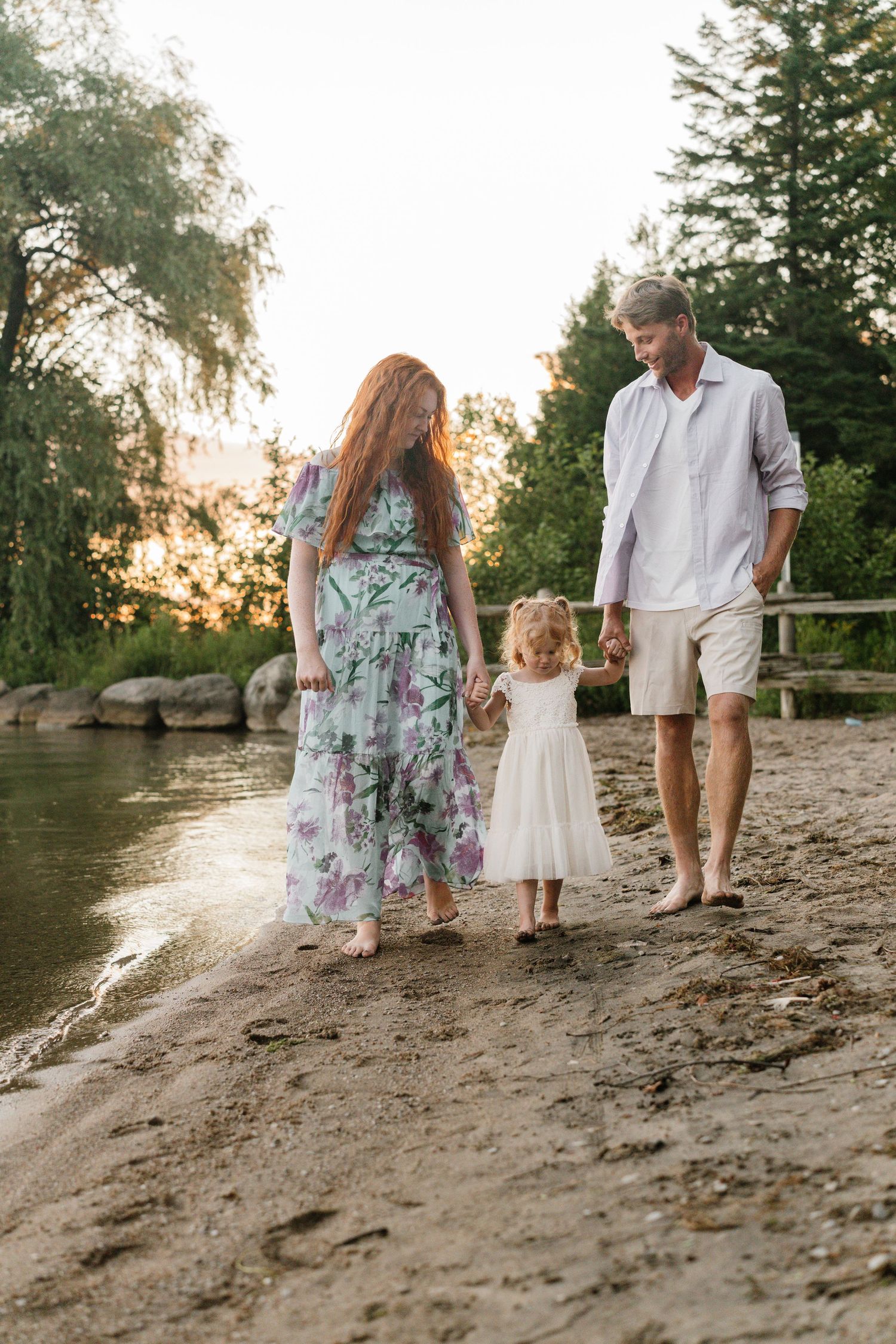 A family of three walks hand-in-hand along a sandy riverbank at sunset, with a little girl in a white dress between her parents.