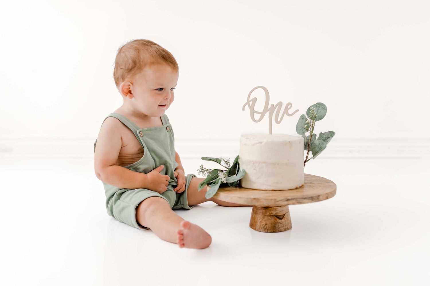 Baby in mint green romper sits on white background with wooden cake stand and eucalyptus sprigs.