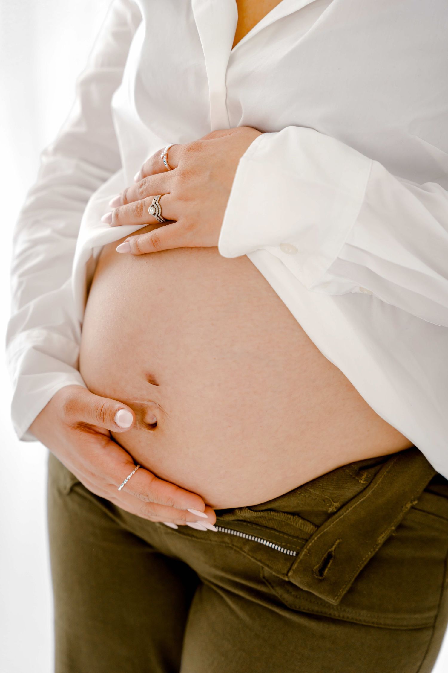 Close up of a pregnant woman's belly in a white shirt and brown pants showing the progression of pregnancy growth.