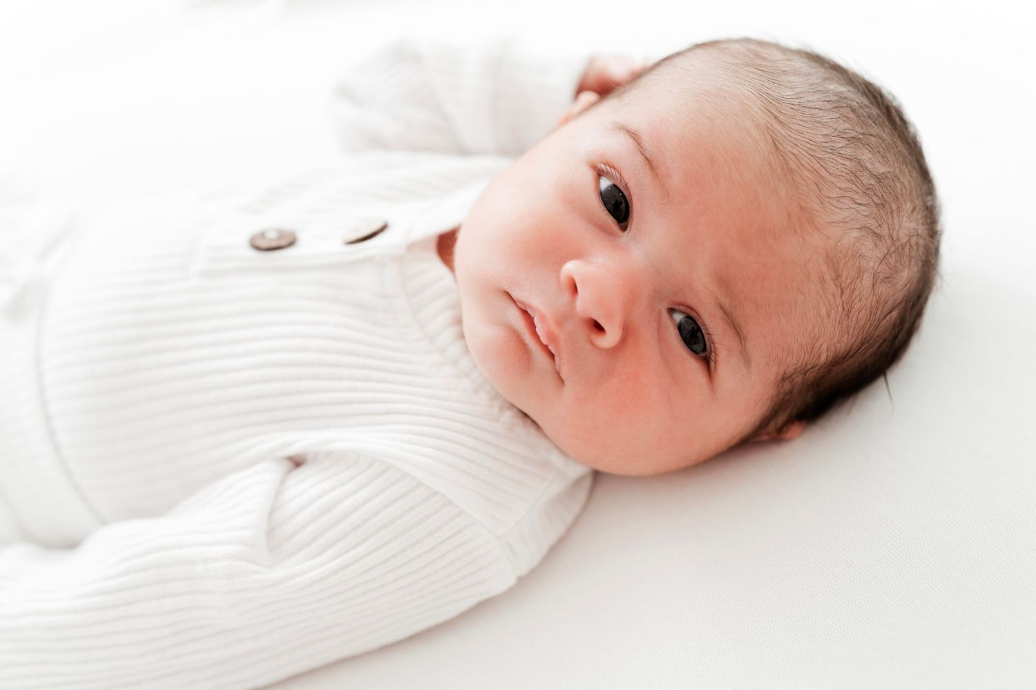 Newborn baby in white outfit lies awake on soft white blanket during studio photoshoot.