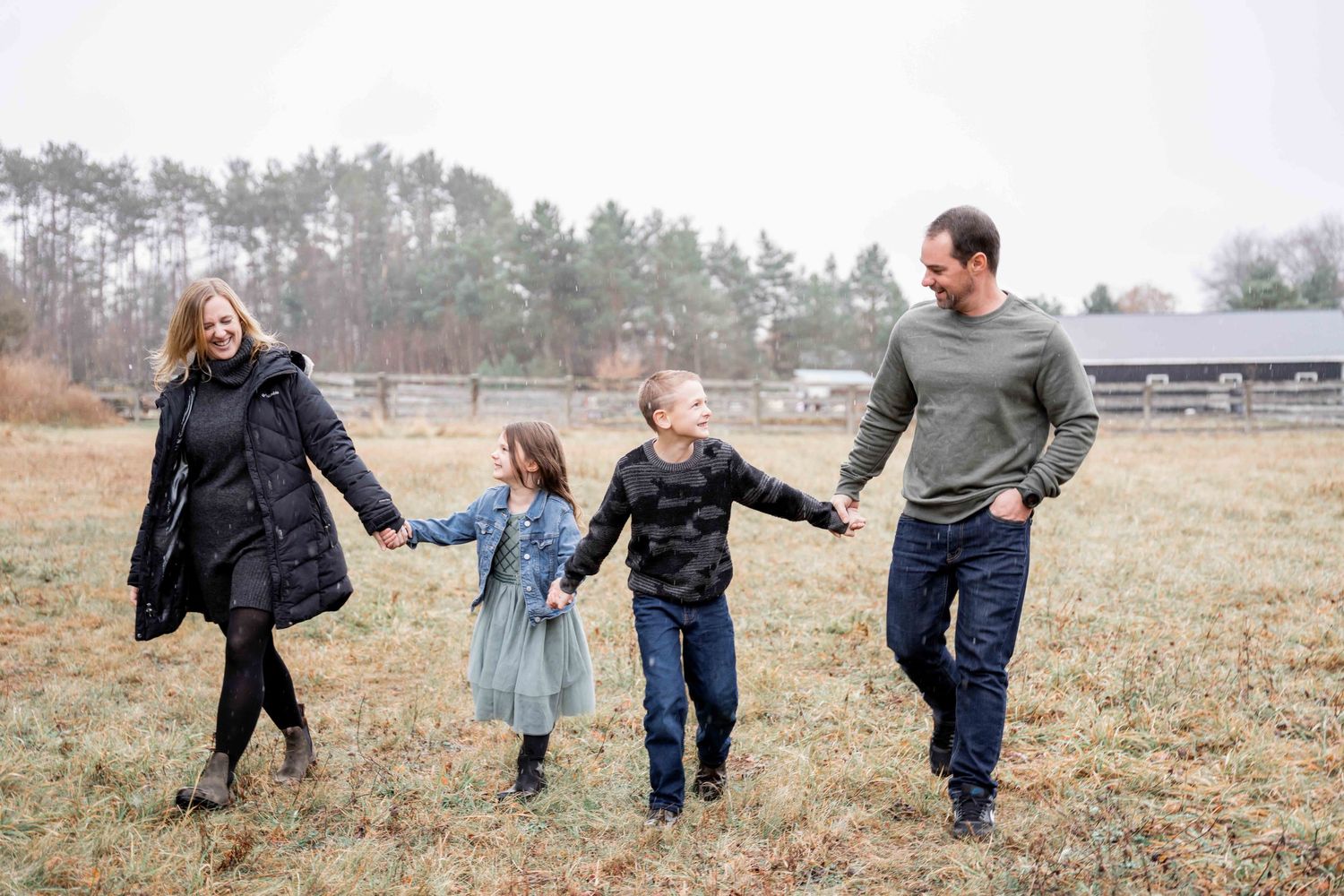 A family walks hand in hand through a misty field with pine trees and a barn in the background.