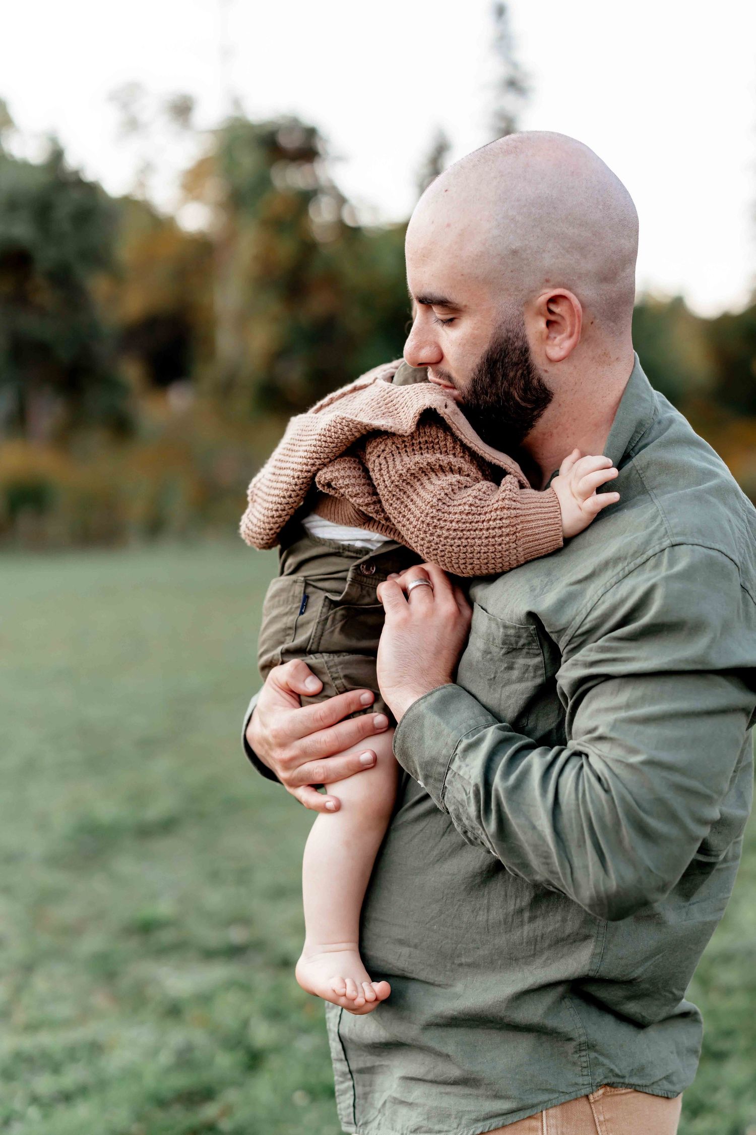 A tender moment captured as a parent in an olive jacket embraces and kisses a baby wearing a brown knit outfit outdoors.