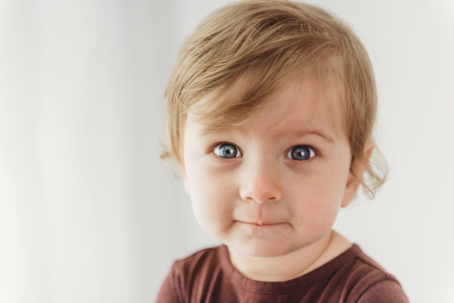 A young toddler in a brown shirt looks at the camera with wide eyes against a white background.