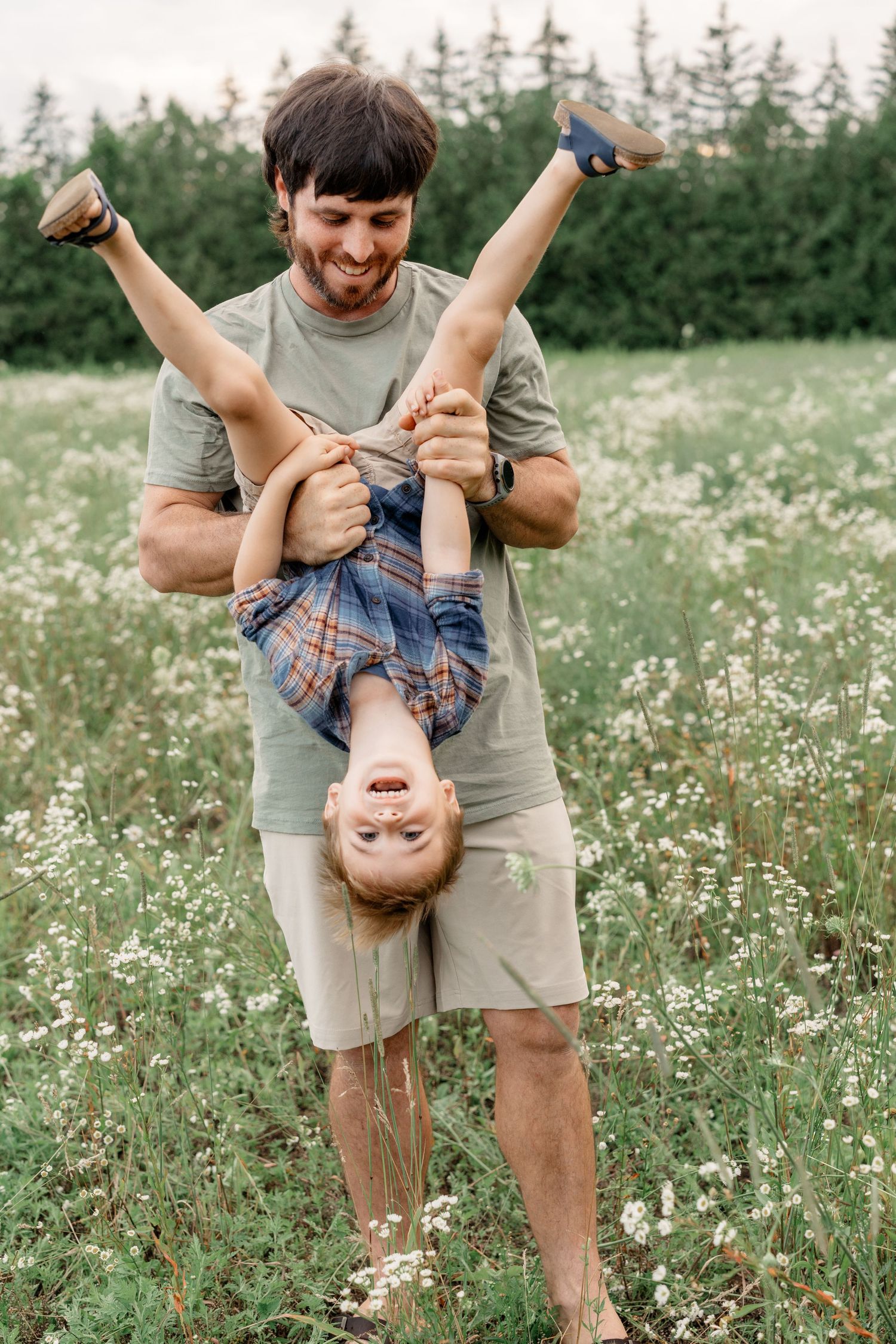 A parent playfully holds a child upside down in a wildflower meadow with evergreen trees in the background.
