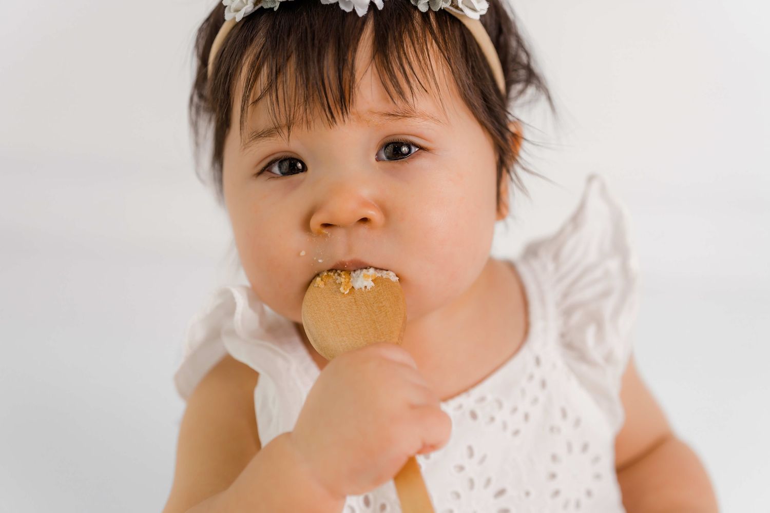 Baby in white lace dress enjoys birthday cake during a cake smash photo session.