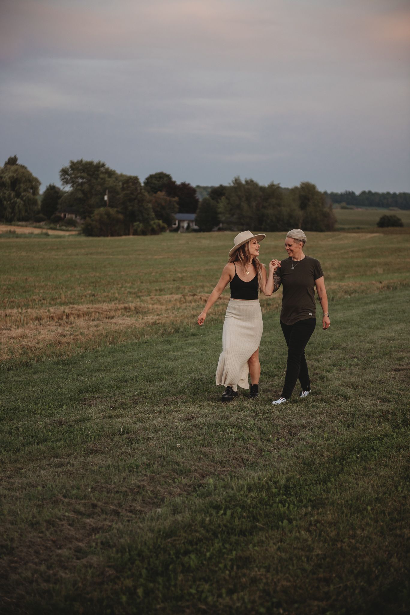 A couple walking together on a grassy field at dusk with trees and buildings in the background.