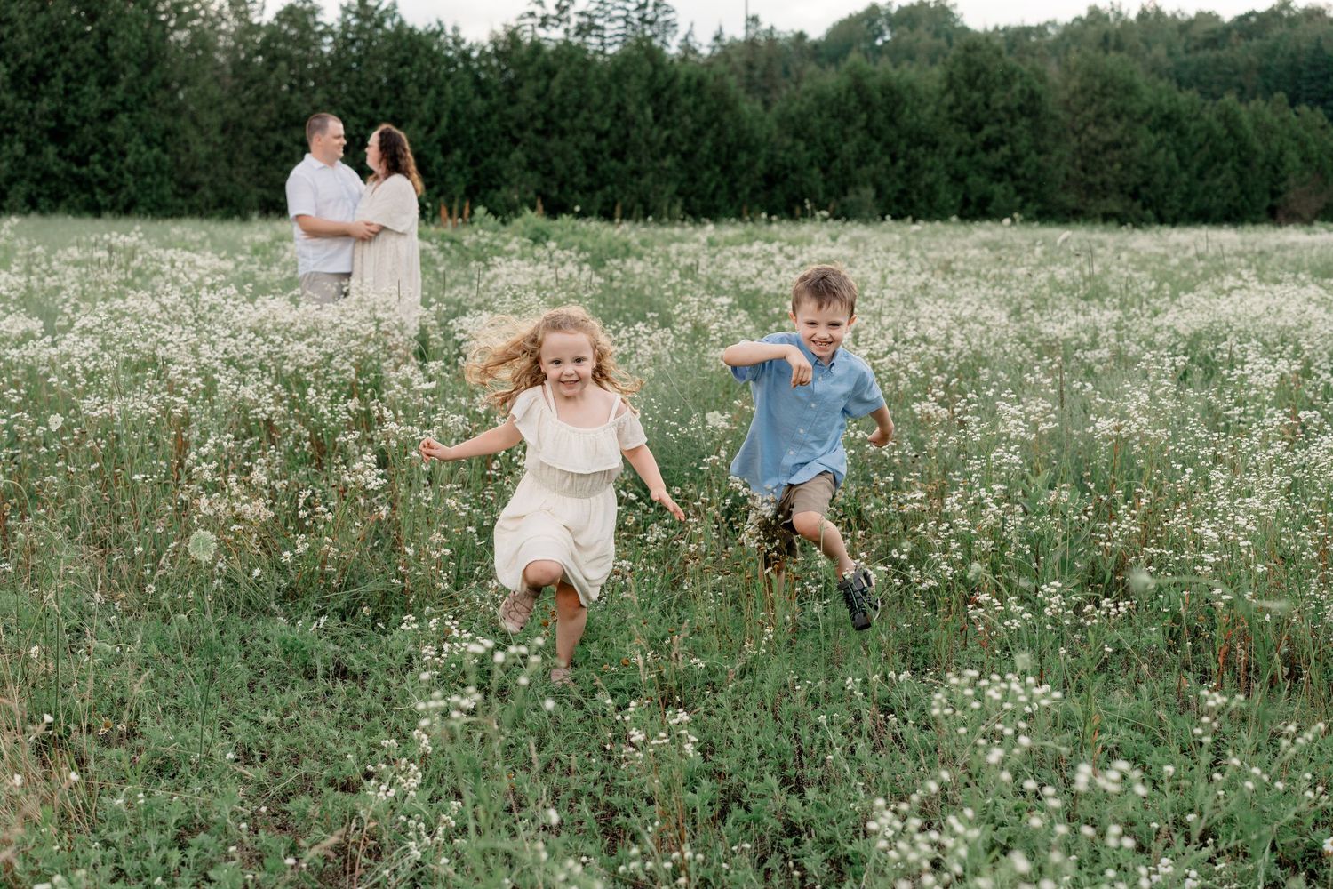 Two children run joyfully through a wildflower meadow while two people stand in the background on a summer day.