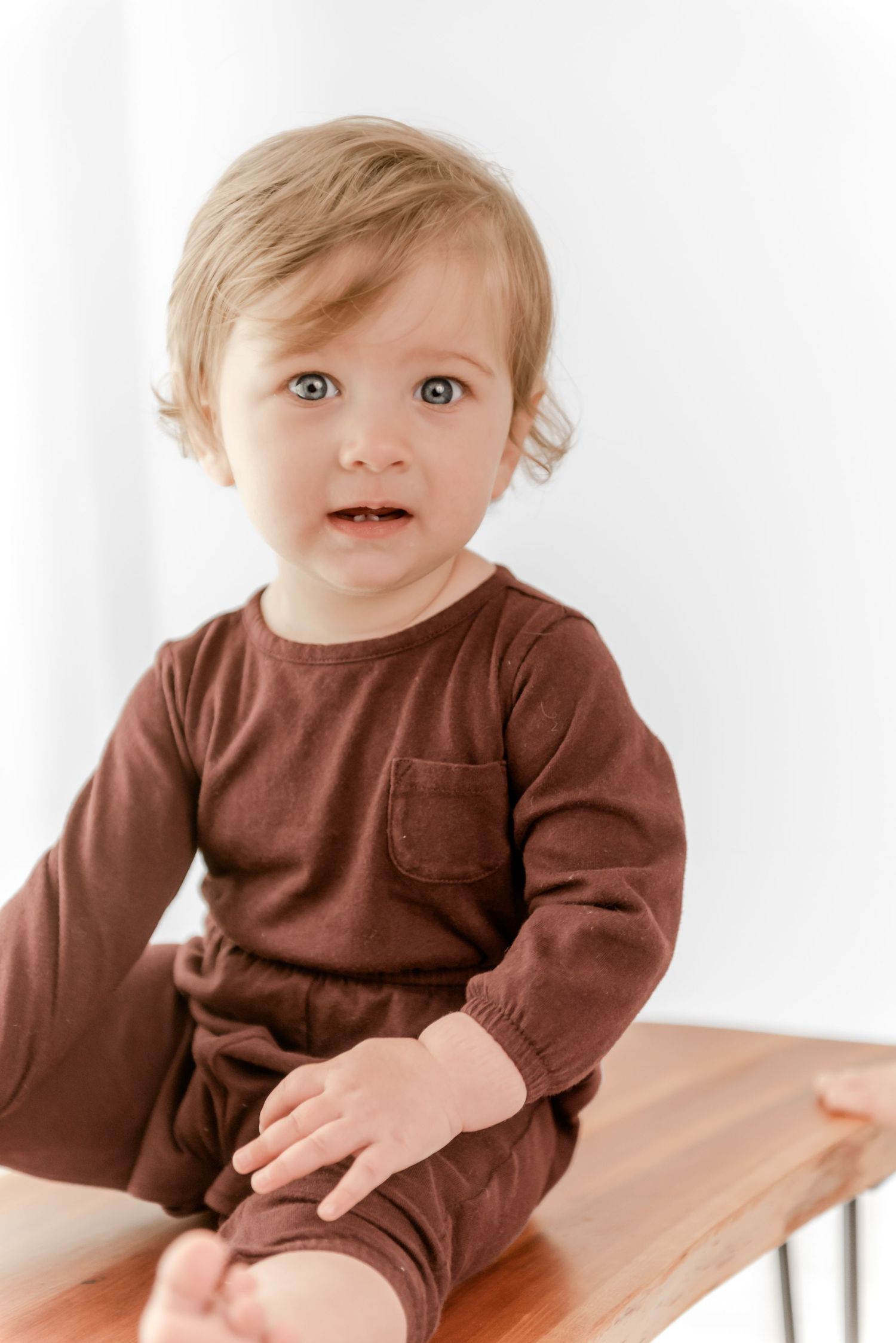 Baby wearing brown long sleeve shirt smiles while sitting on wooden table in bright white room.