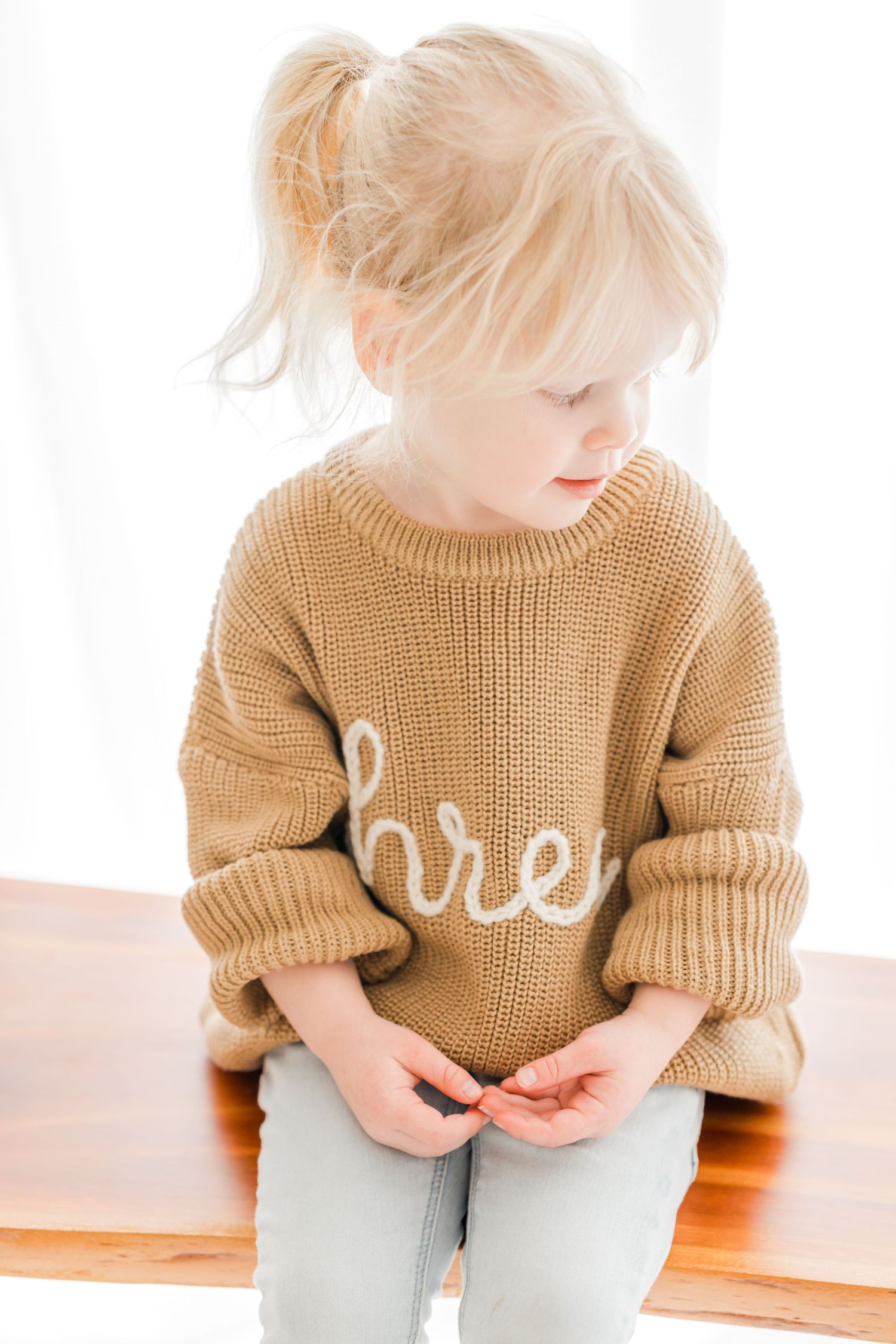 A child in a tan knit sweater sits on a light surface in a bright, minimalist setting.