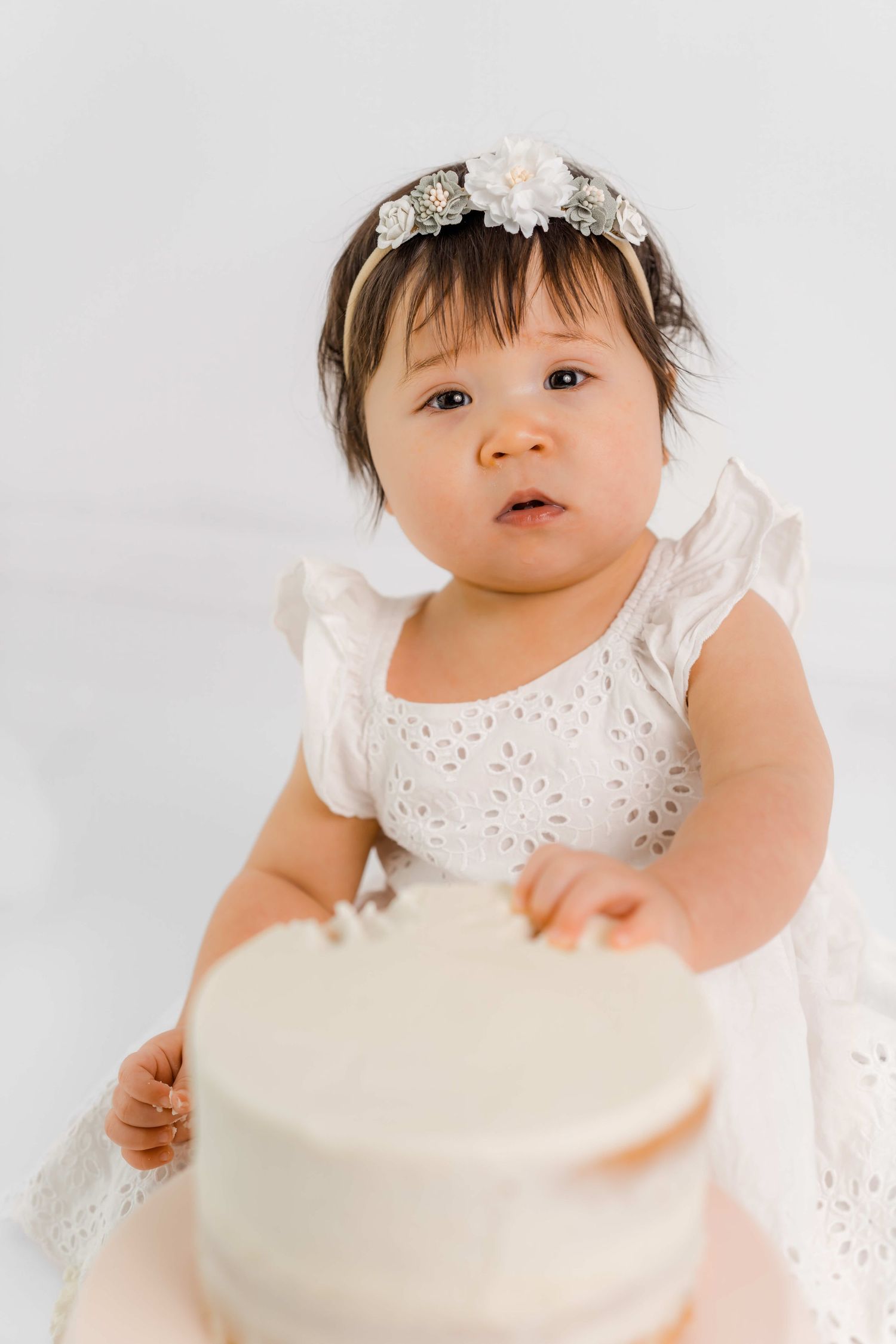 Baby in white eyelet dress and floral headband reaching toward a cream colored cake on white background.