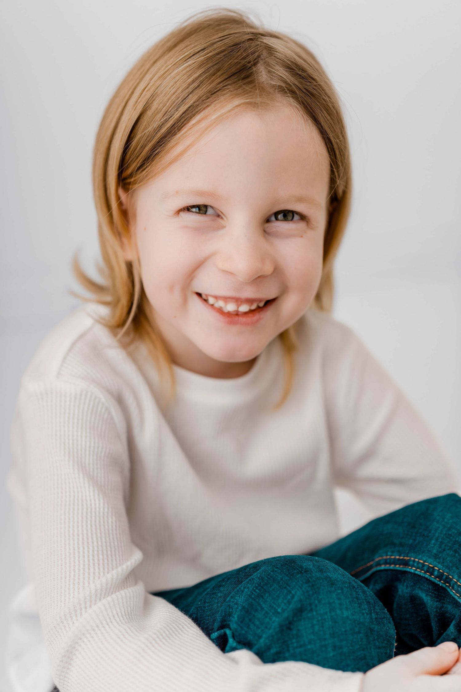 Child wearing white sweater and jeans smiles warmly against light background in studio portraits.