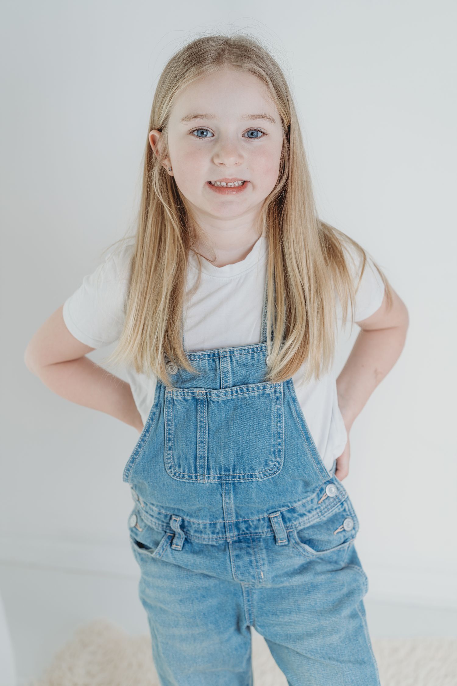 A young child in denim overalls and white t-shirt poses playfully against a light background.