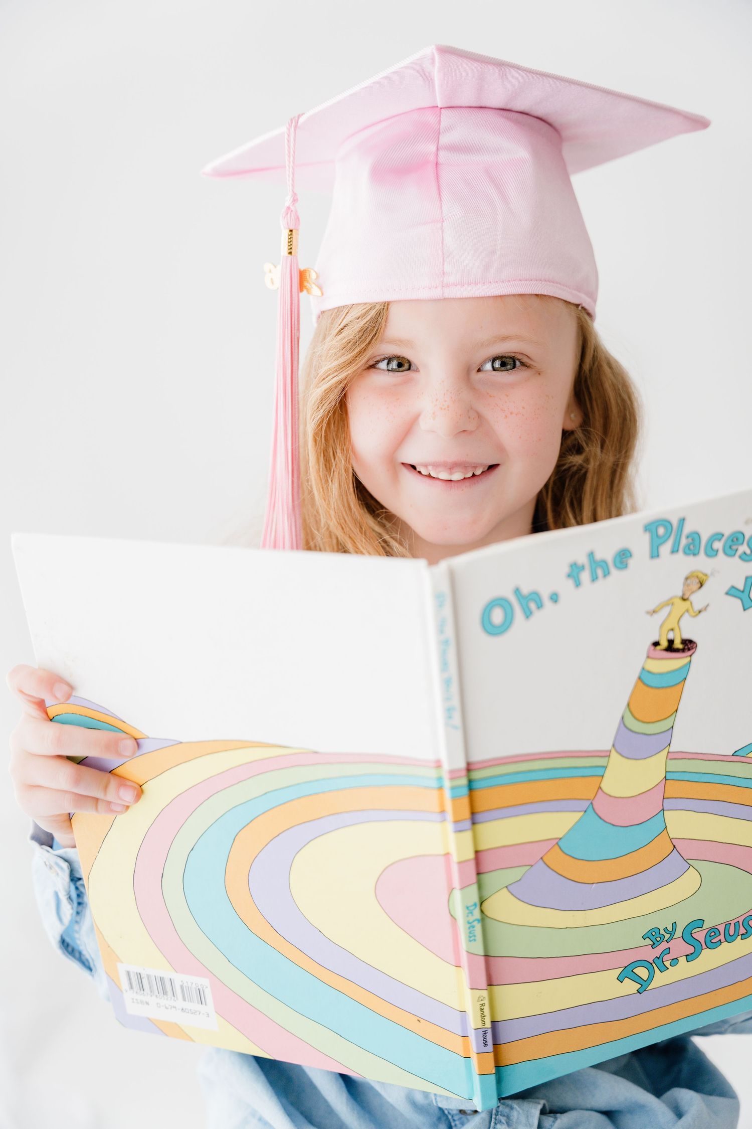 A smiling girl in a pink graduation cap reads Dr. Seuss Oh the Places Youll Go book.