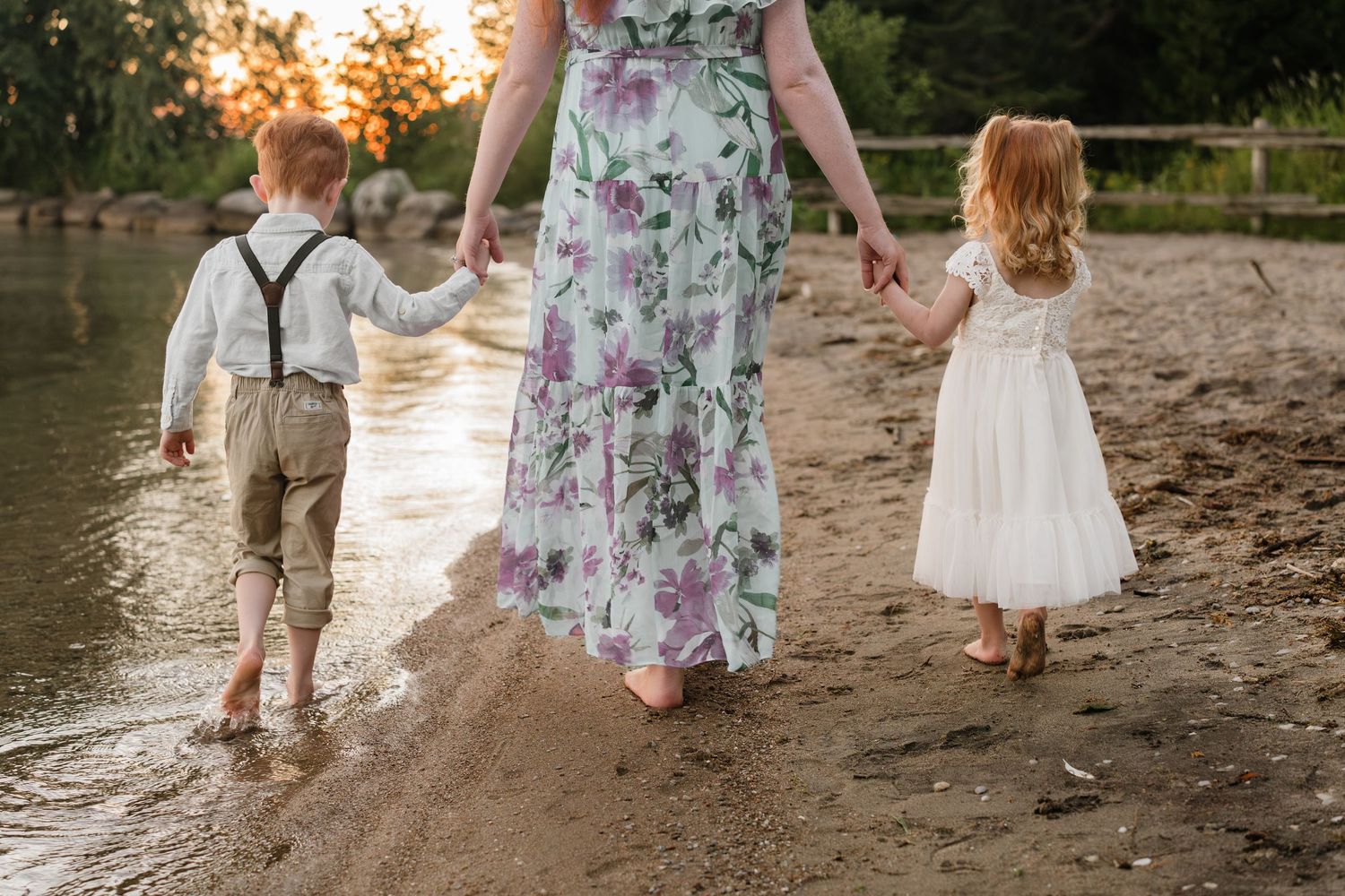 A mother walks barefoot on a sandy beach at sunset, holding hands with her young son and daughter.