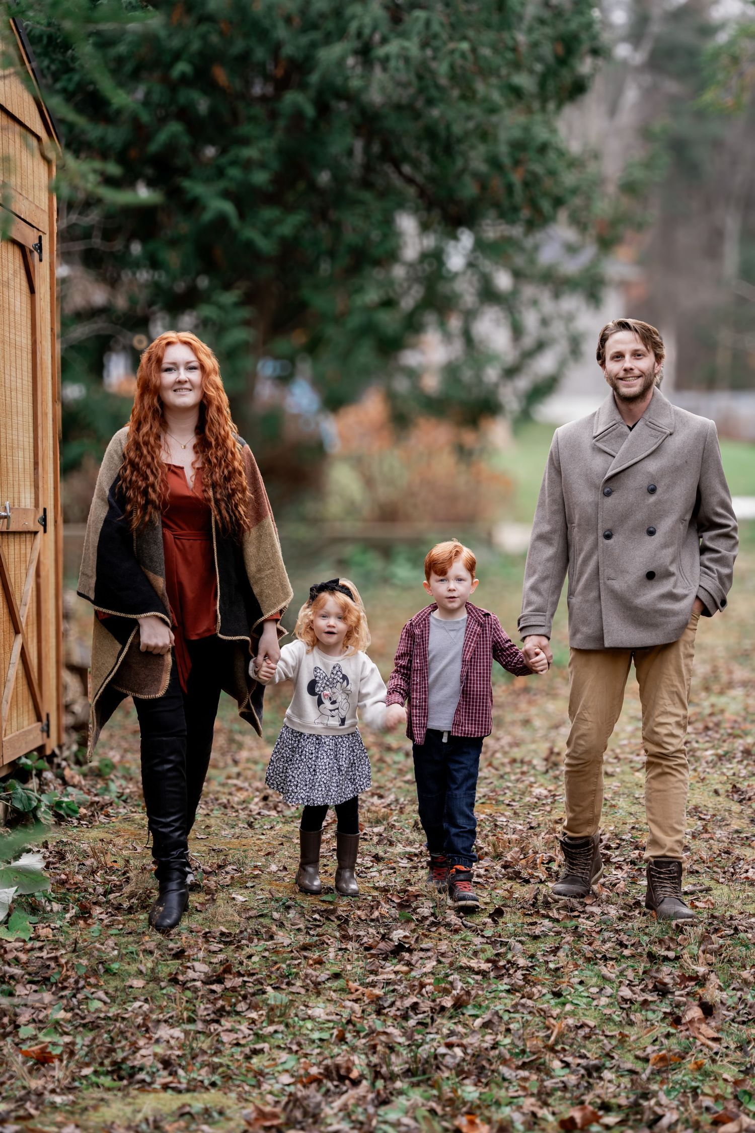 A family walks together through fallen autumn leaves in a garden setting wearing coordinated casual fall outfits.