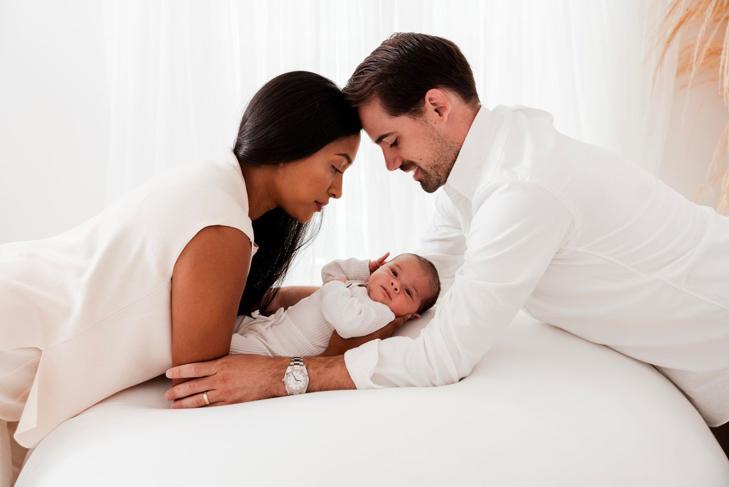 Family shares intimate moments with newborn baby on white bedding in natural light.