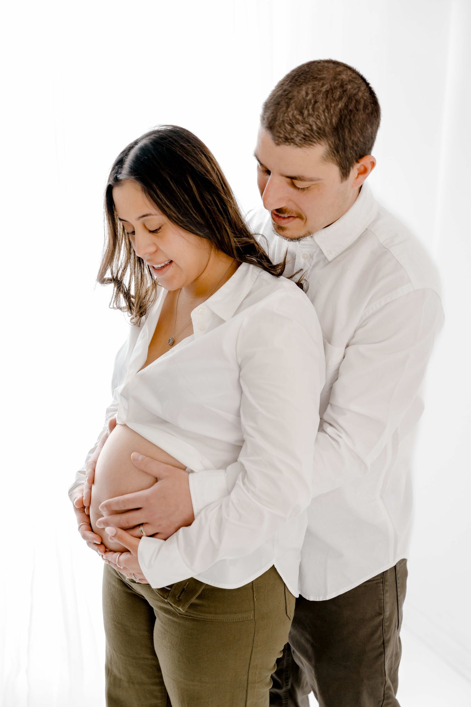 Couple in white shirts and olive pants embrace tenderly during maternity photoshoot against white background.