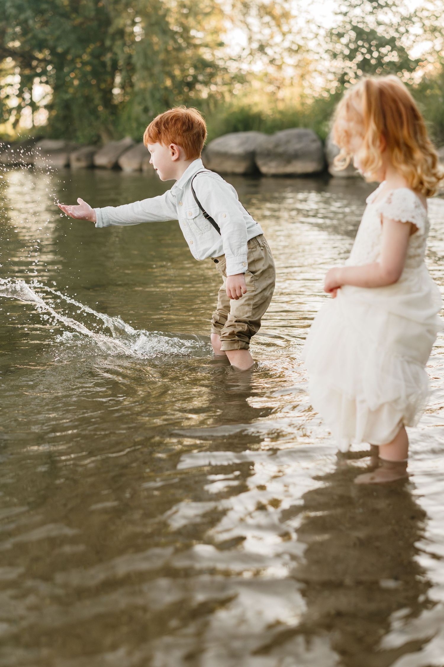Two redhead children playing in shallow river water, boy splashing in suspenders and girl in white dress.