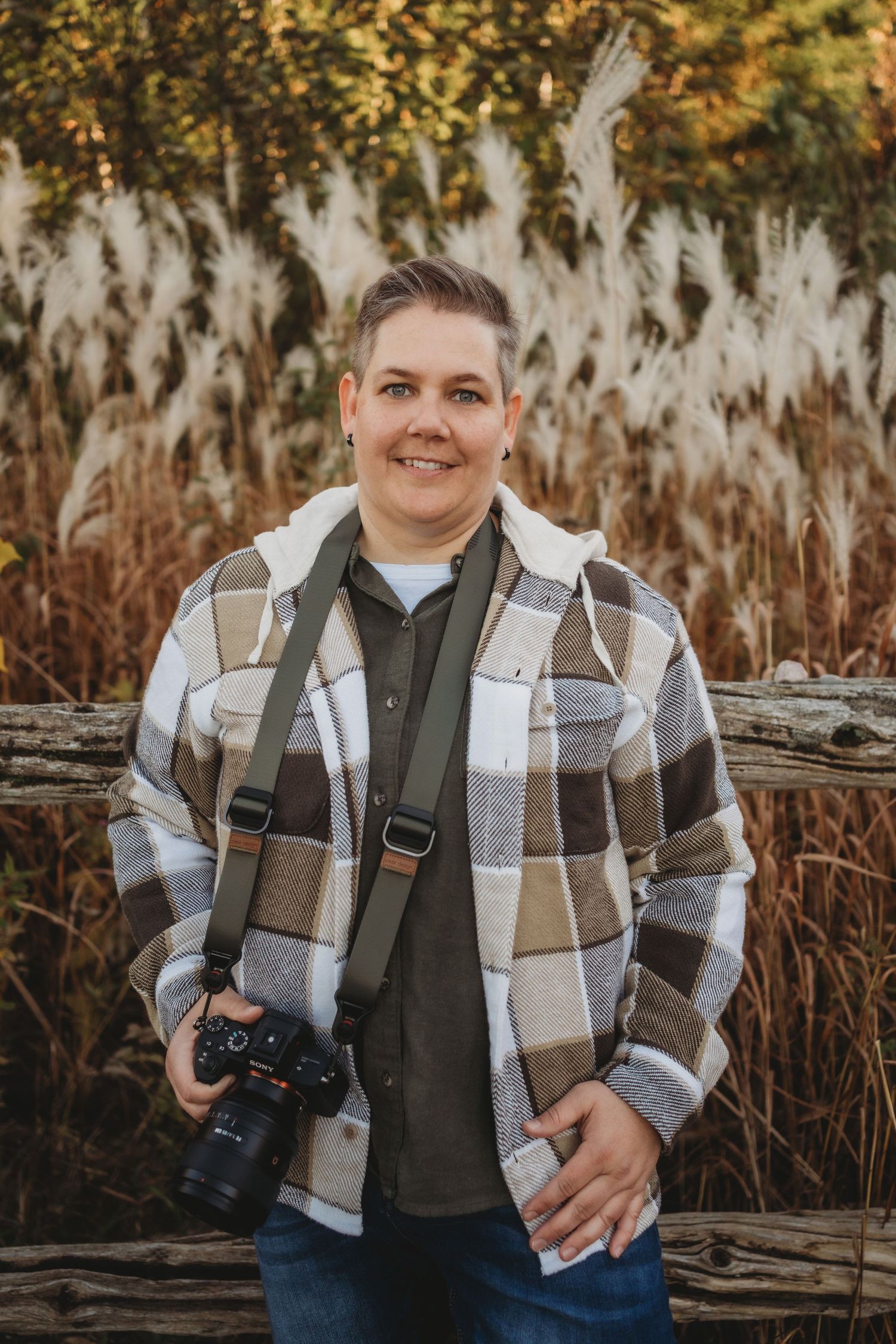 A photographer in a plaid jacket and olive vest poses outdoors against a rustic wooden fence and autumn foliage.