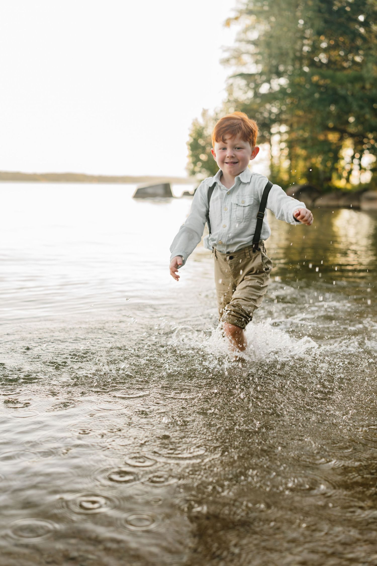 Joyful redhead boy splashing through shallow lake water wearing suspenders and khaki pants on a sunny day.