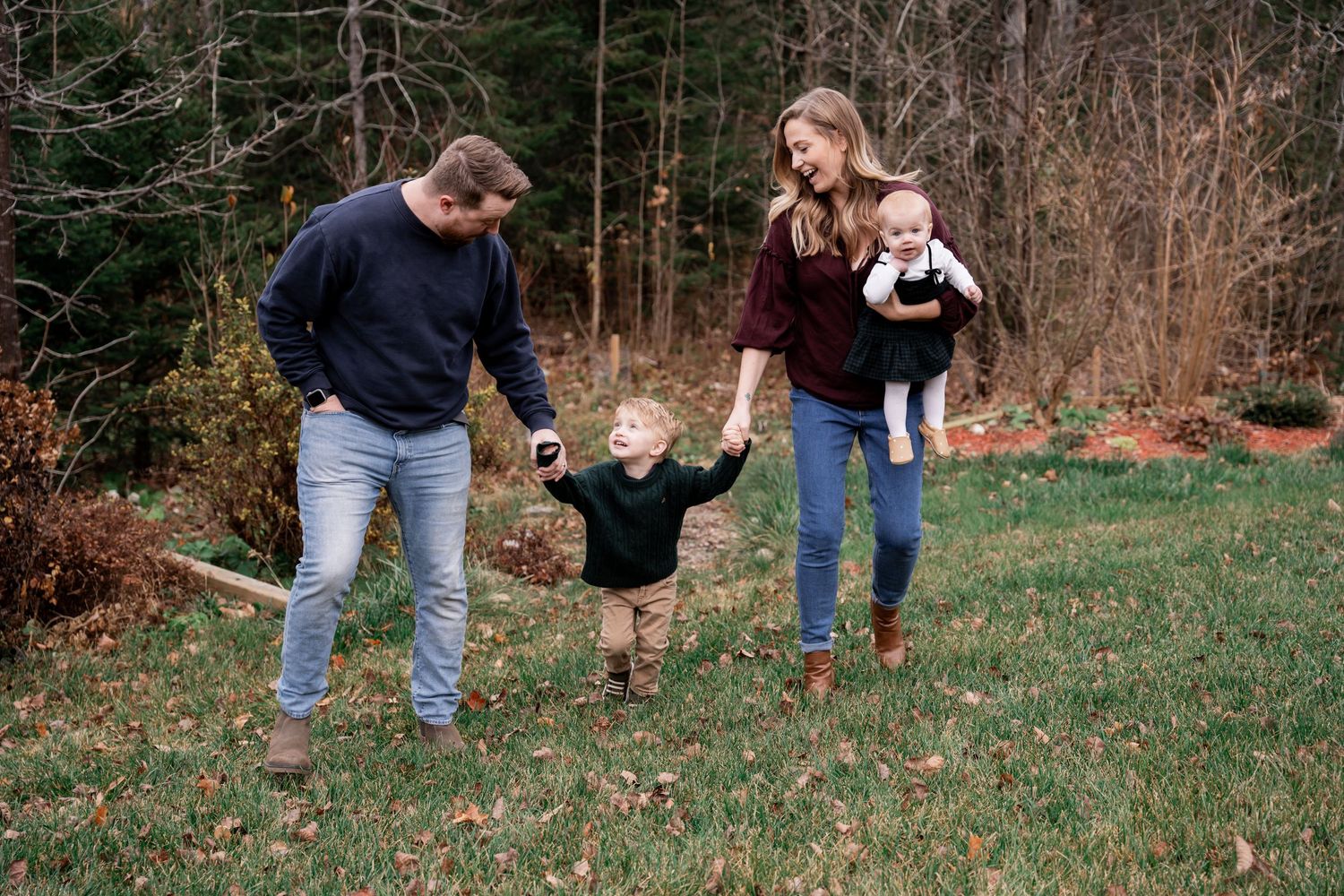 A family holds hands and walks together on a grassy hill with evergreen trees in the background during autumn.