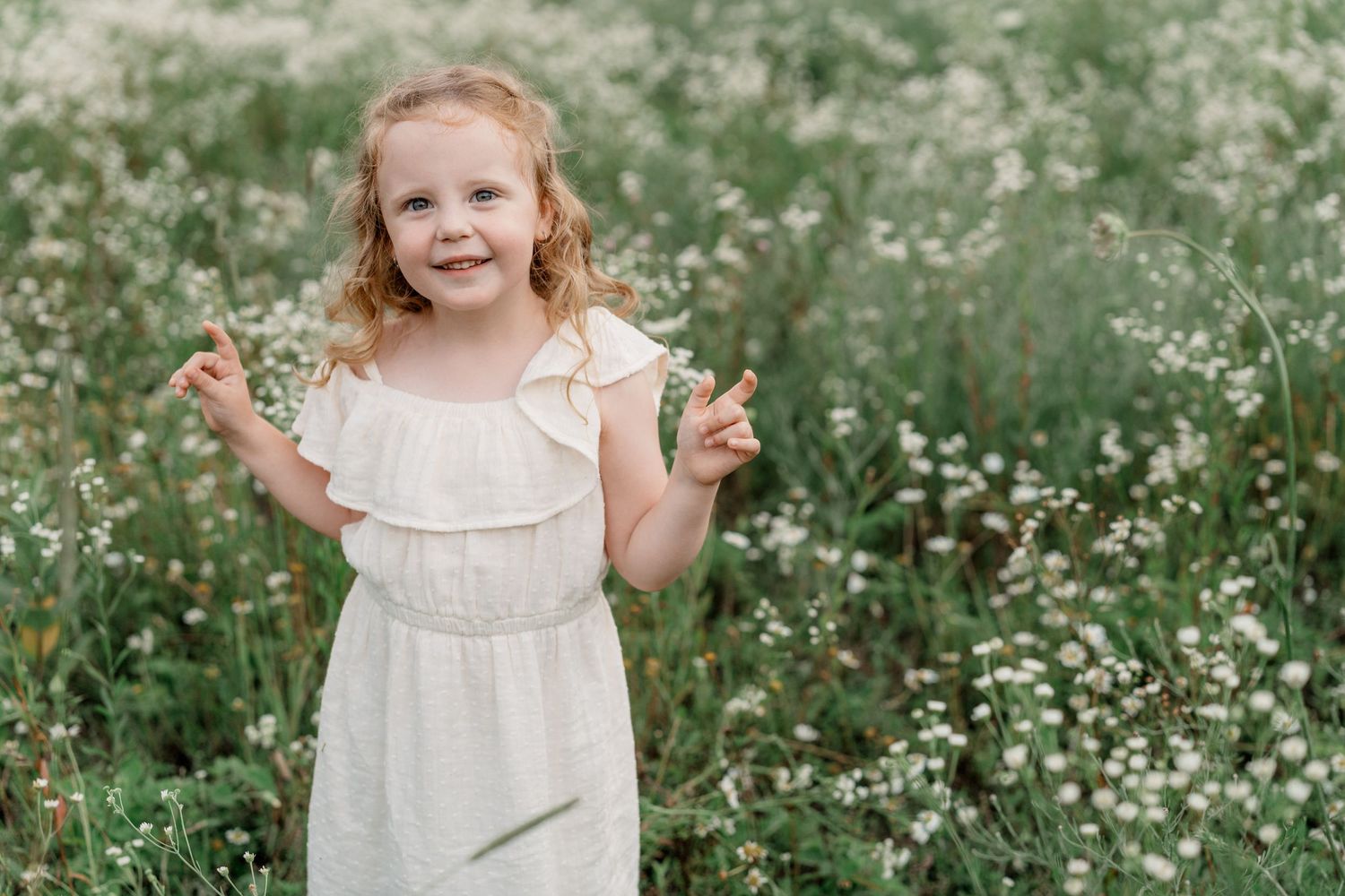 Child in white dress plays joyfully among blooming wildflowers in a green summer meadow.