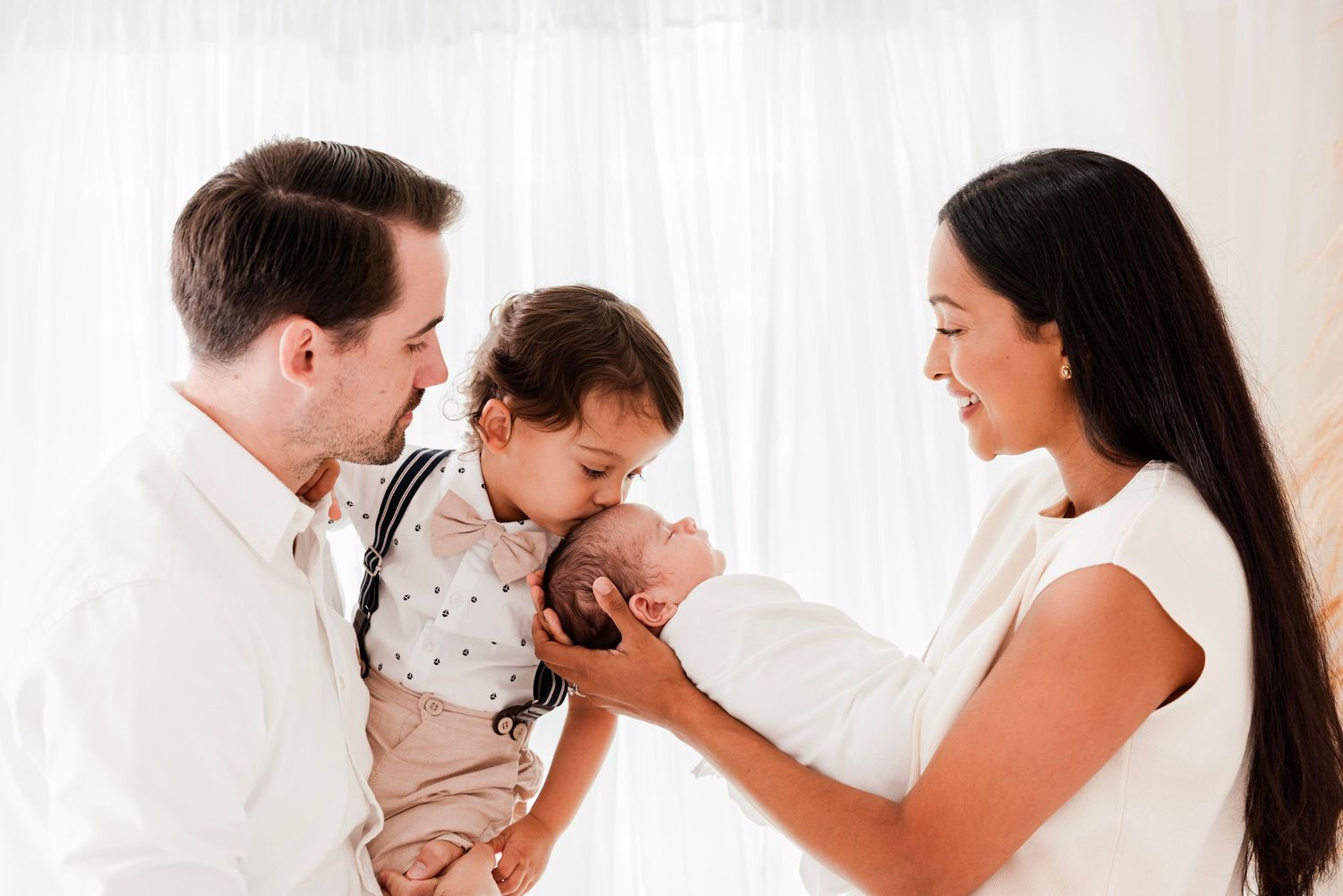 Parents share tender moments with their newborn baby and toddler in bright white studio setting.