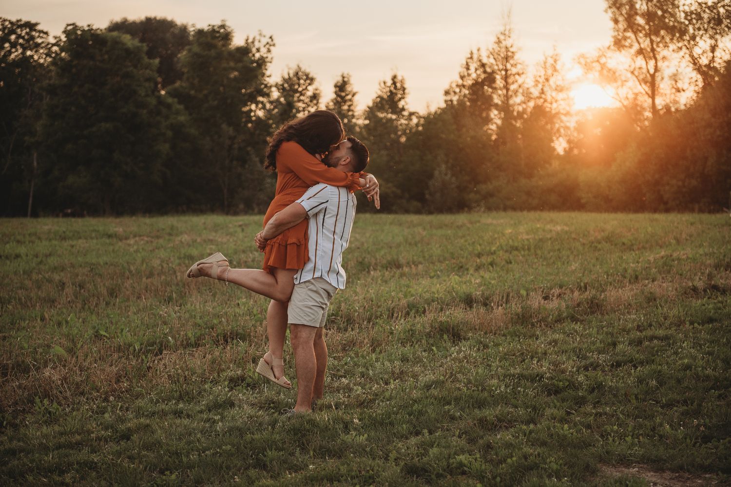 A couple embraces in a golden meadow at sunset while wearing casual summer attire.