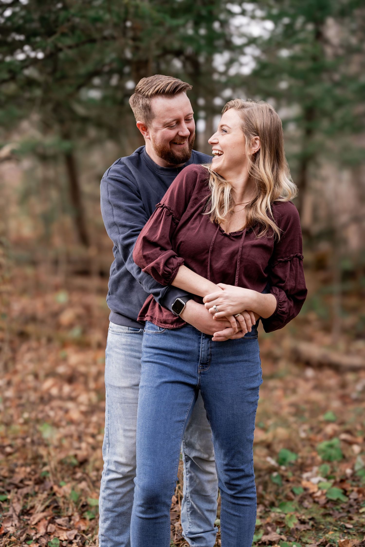 A couple shares joyful laughter while embracing in a wooded autumn setting with fallen leaves on the ground.