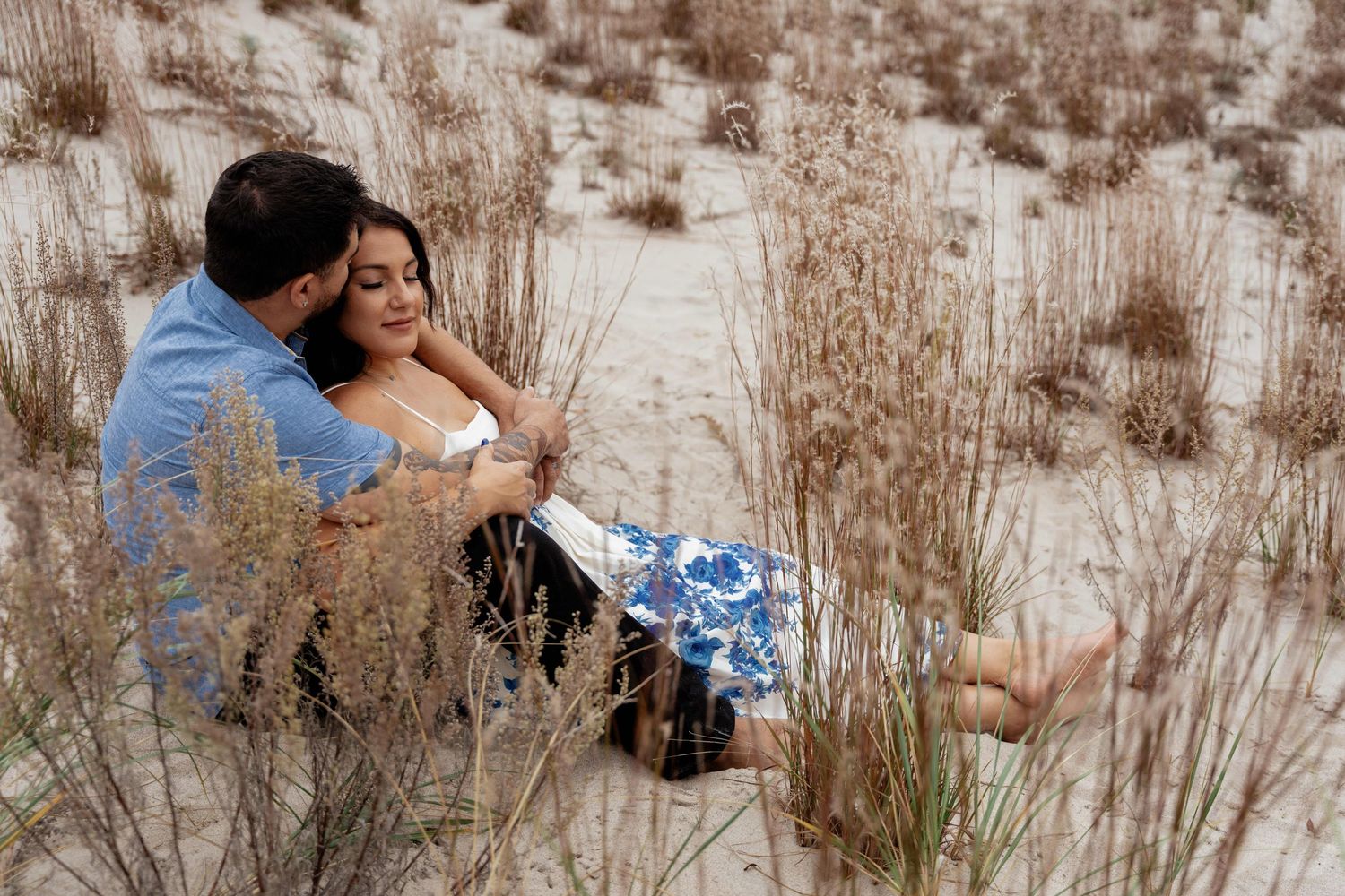 A couple embraces in a rustic outdoor setting with tall dried grass and natural scenery in the background.