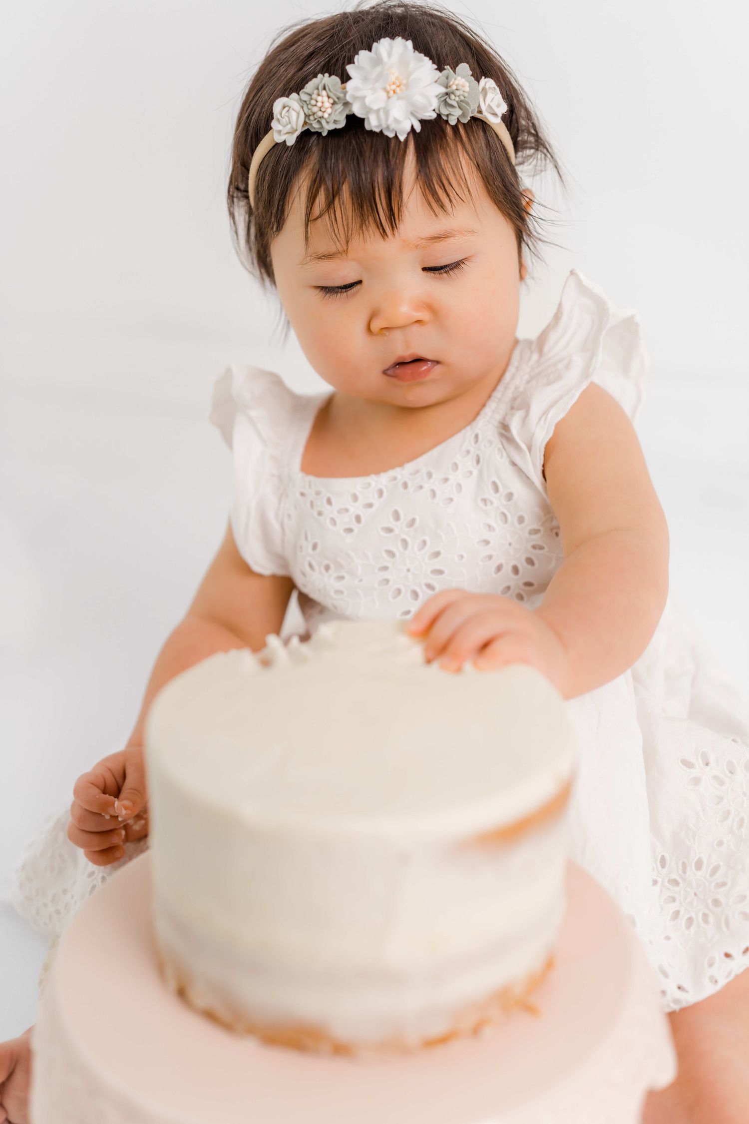 Baby in white eyelet dress explores white frosted cake during cake smash photo session against light background.