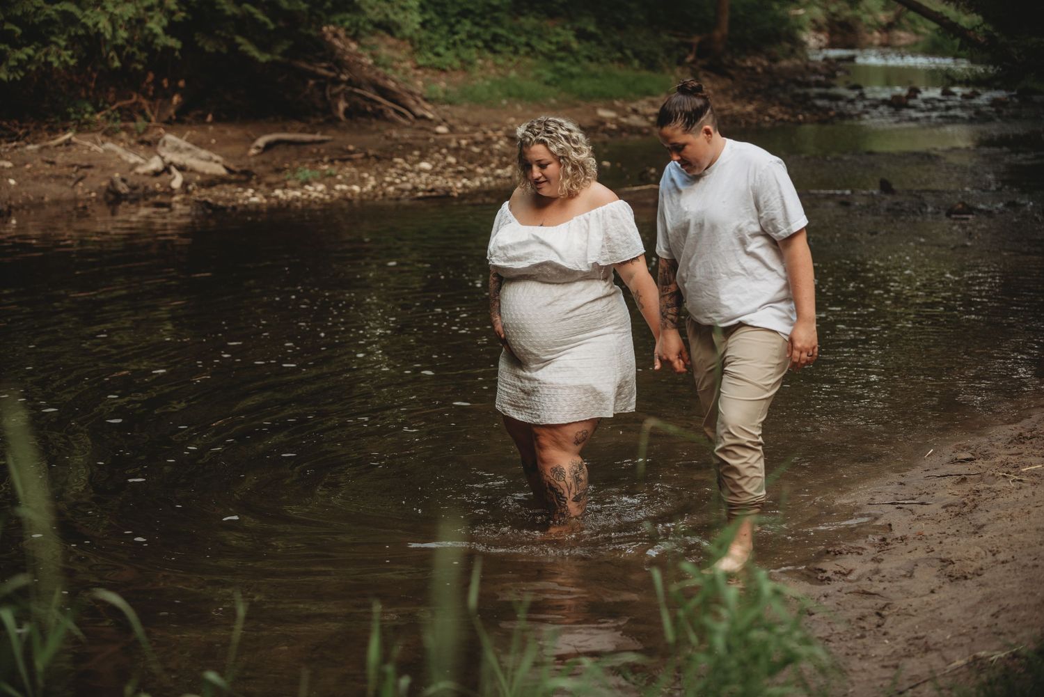 A couple walks hand in hand along a peaceful creek bank surrounded by lush greenery and natural scenery.
