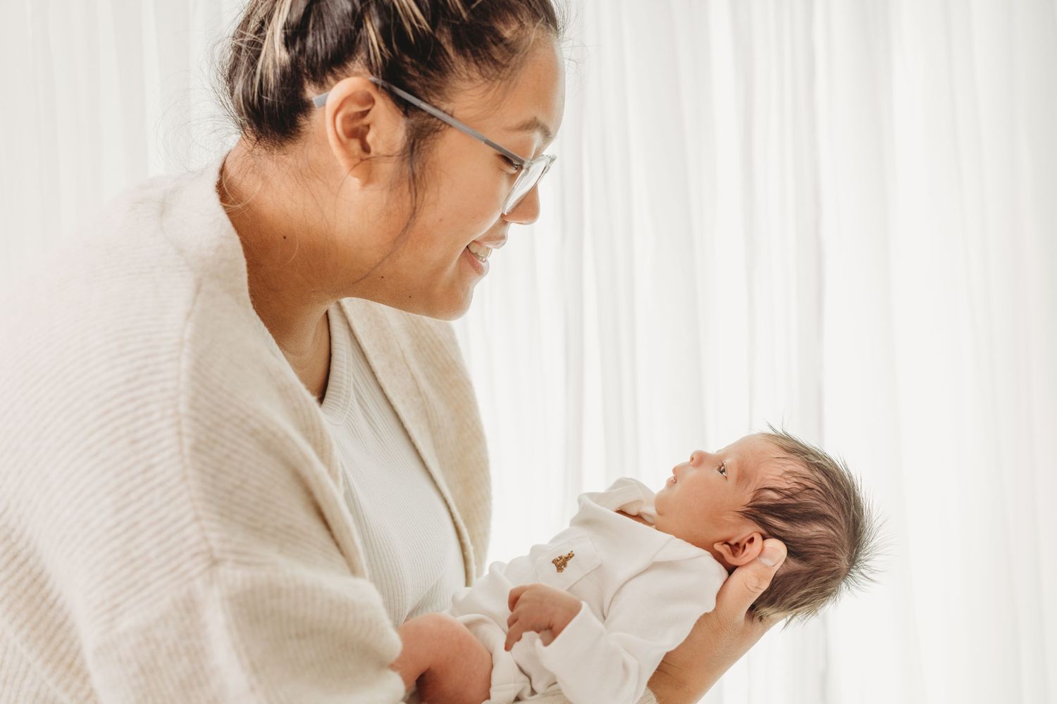 A mother in a white sweater lovingly cradles and gazes at her newborn baby in a bright, peaceful nursery setting.